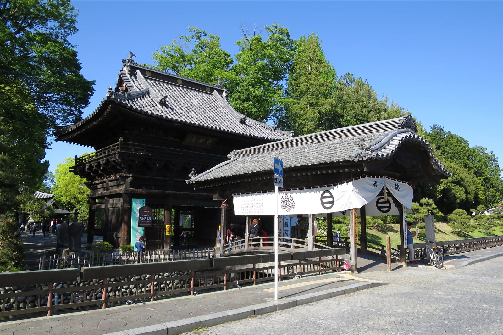 晴れた空の下、堀に架かる朱色の太鼓橋と、その奥に建つ鑁阿寺の楼門