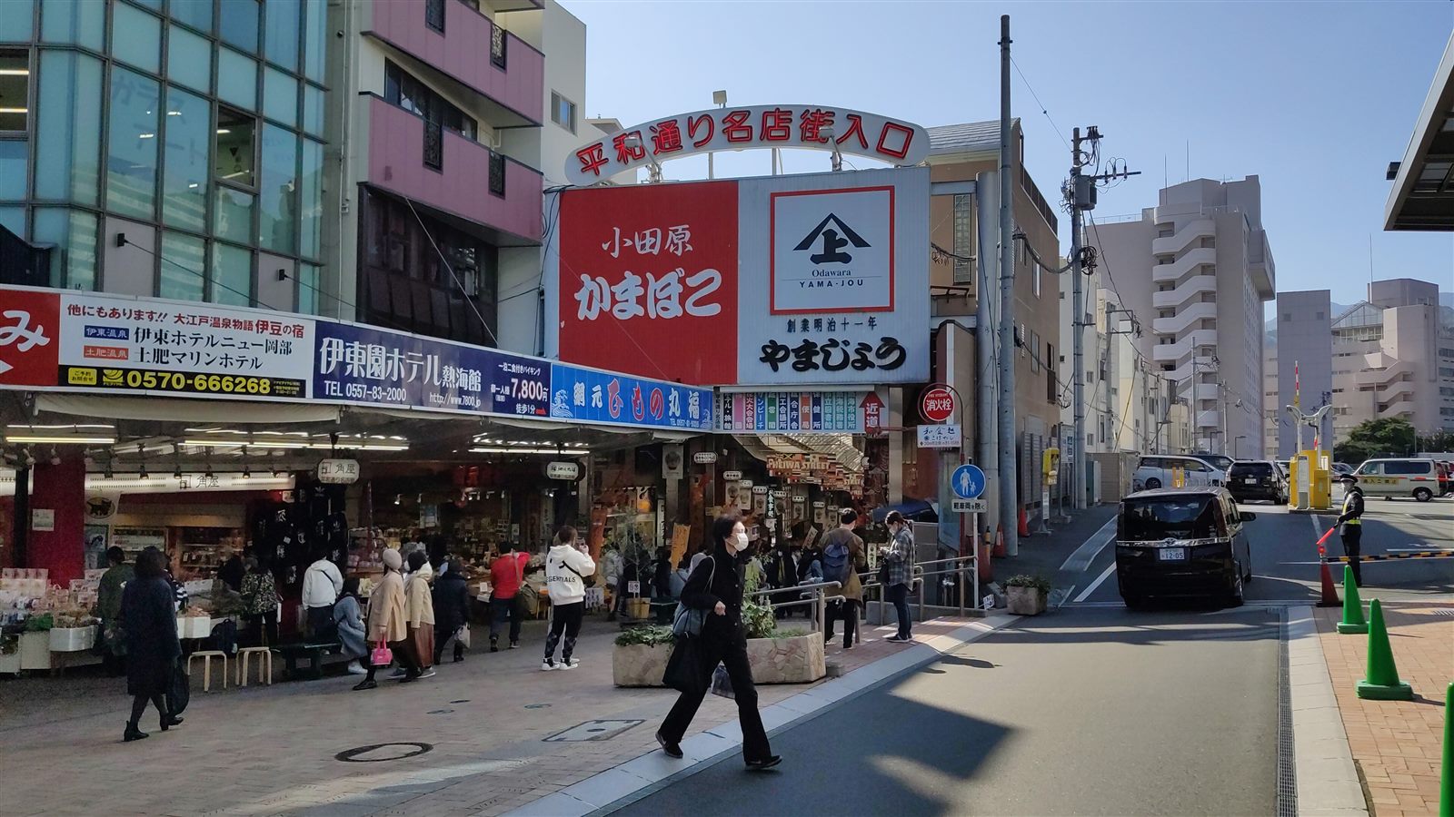 熱海駅前の平和通り名店街の入口。アーチ形の看板の下に、かまぼこ店の大きな看板とみやげ物の並ぶ店先があり、歩道を人々が行き交っている
