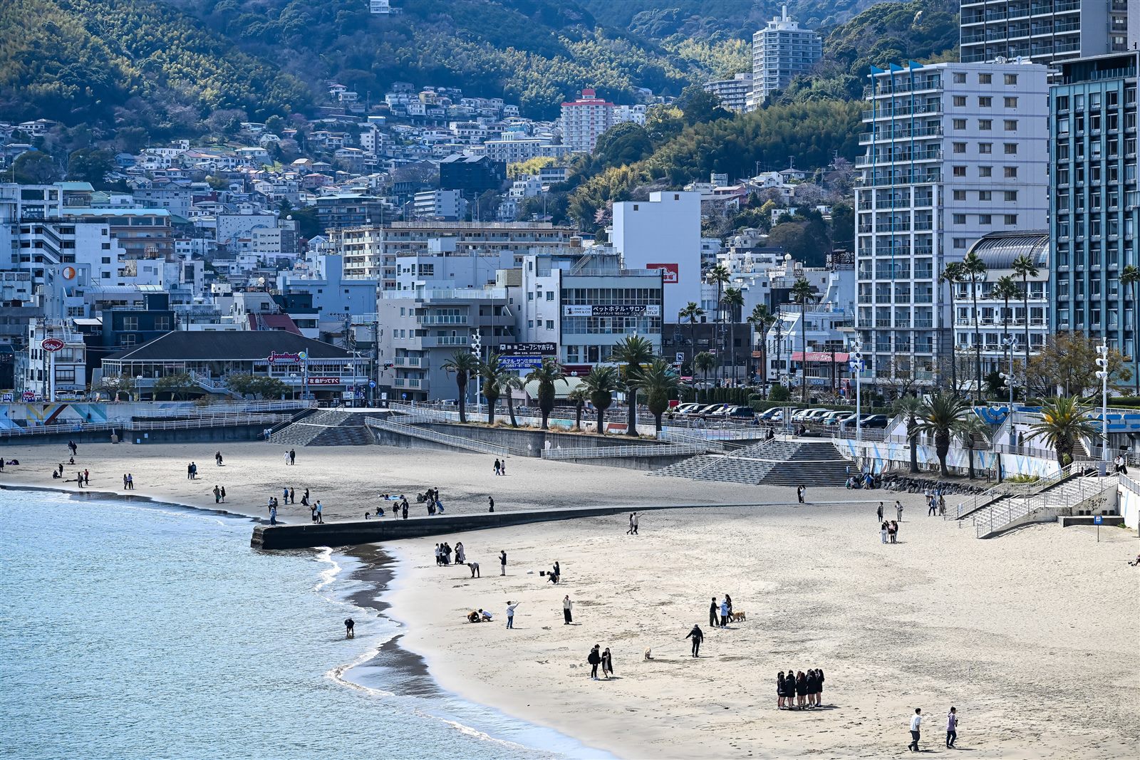 晴れた日の熱海サンビーチ。弧を描く砂浜を人々が散歩し、奥にはヤシの木の並ぶ海沿いの道と、山の斜面を埋めるホテルや住宅が見える