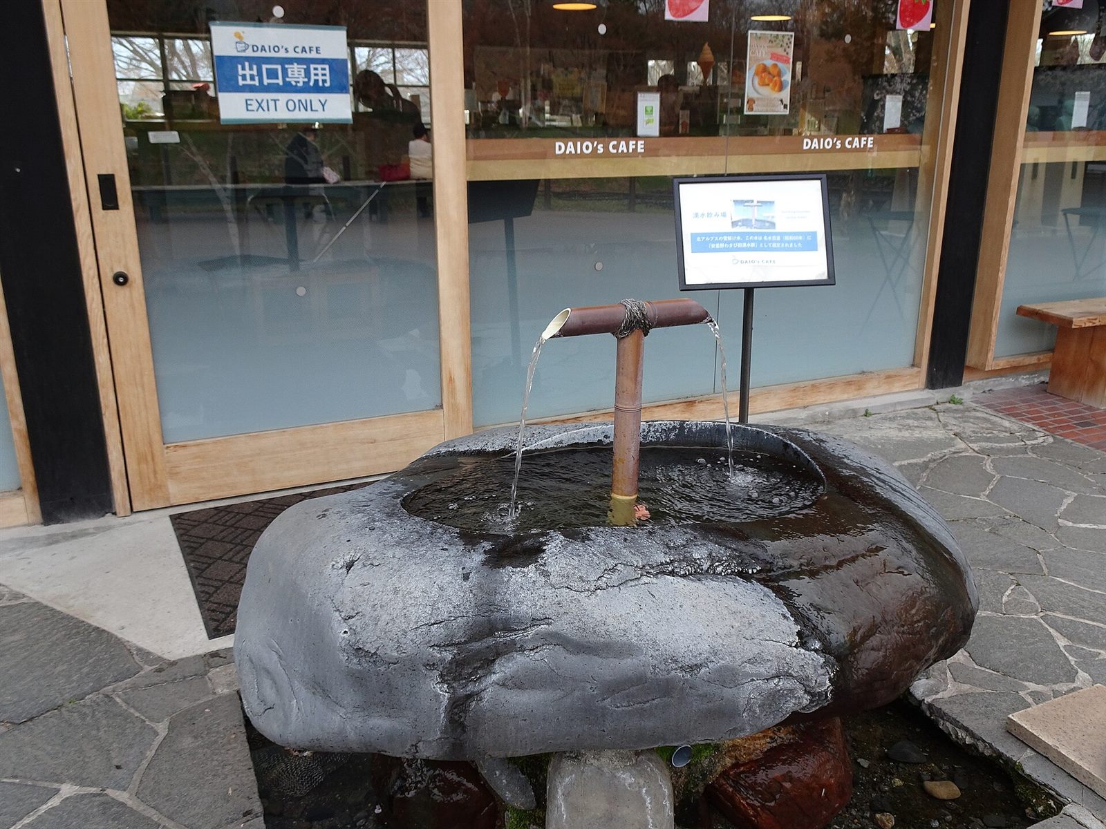 大きな石の水盤に、竹の筒から湧き水が注ぎ、後ろのガラス越しにカフェの店内が見える