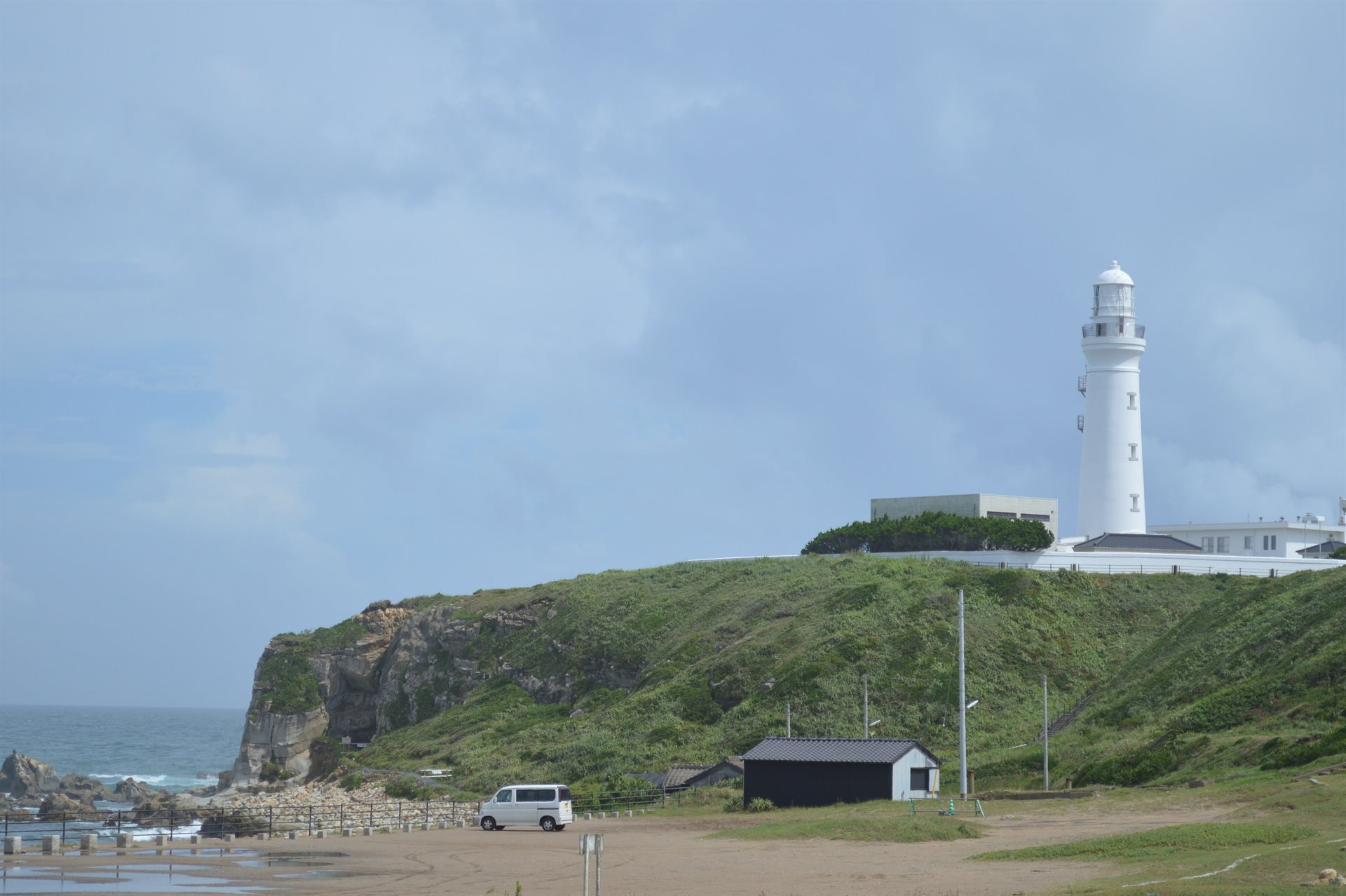 浜から見上げた犬吠埼灯台。緑に覆われた崖の上に白い灯台が立ち、左手に岩の磯と波、手前に砂地が広がっている