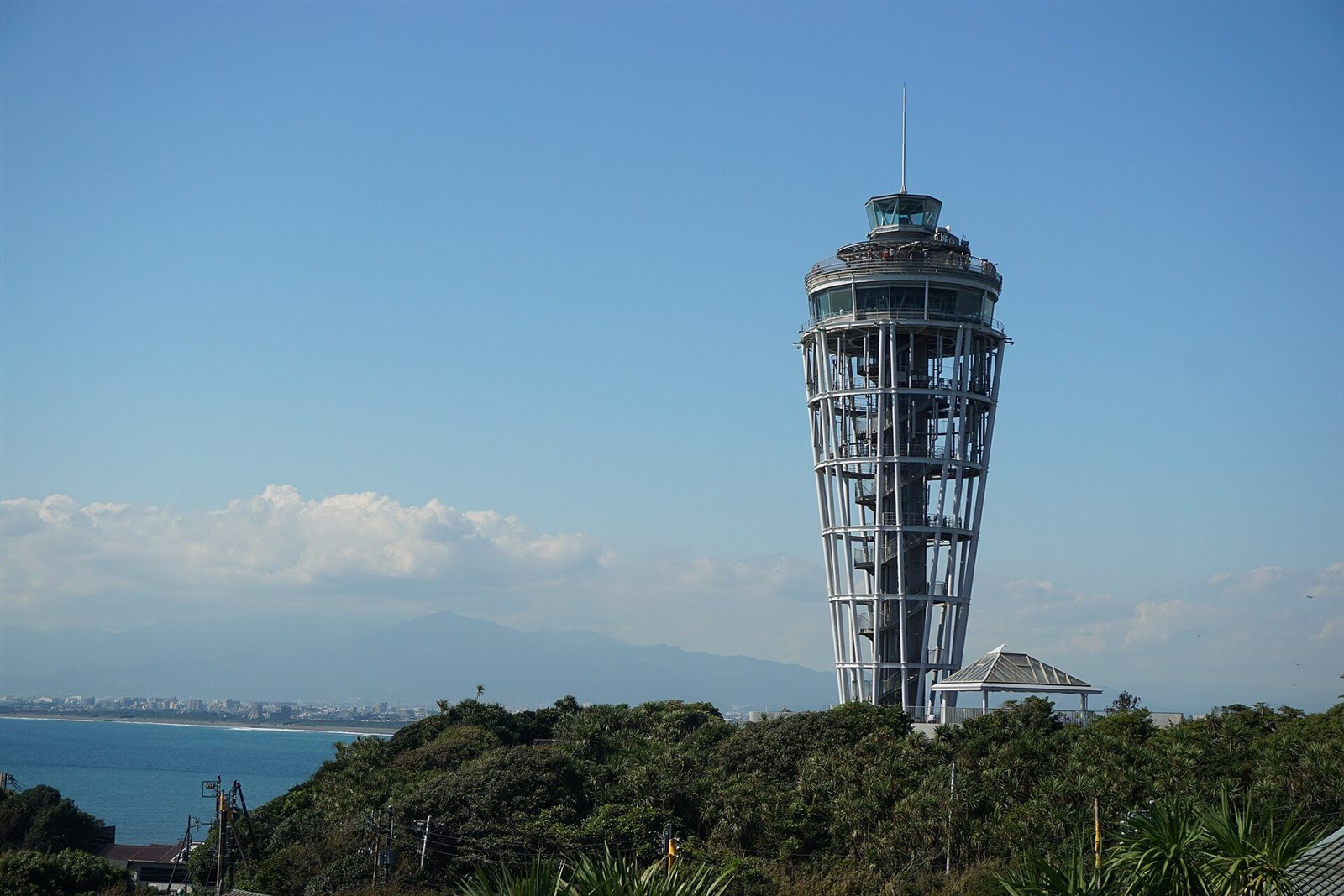 青空の下、島の緑の上に立つ白い展望灯台。左奥に海と対岸の街並み、遠くに山が見える