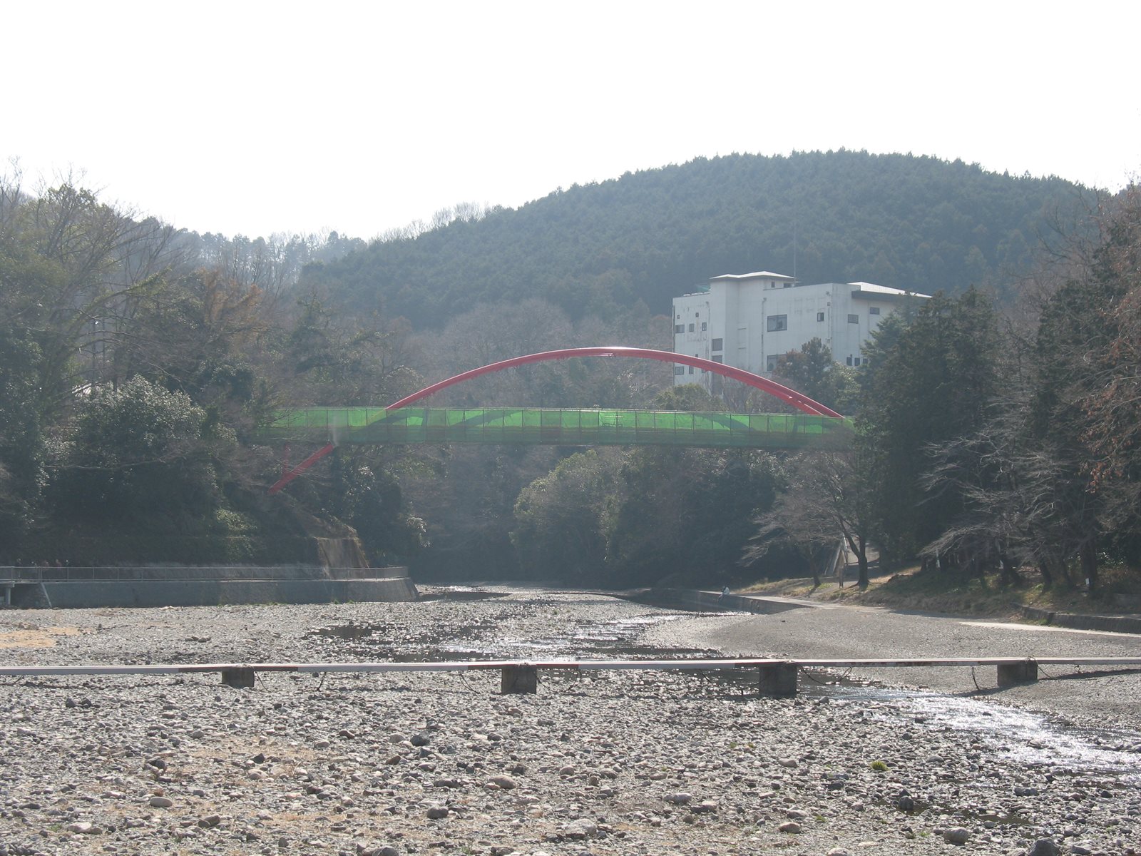 飯能河原から下流を見た入間川。手前に石の広がる河原と細い流れがあり、その先に赤いアーチの割岩橋が架かる。奥に木の茂る山が重なっている