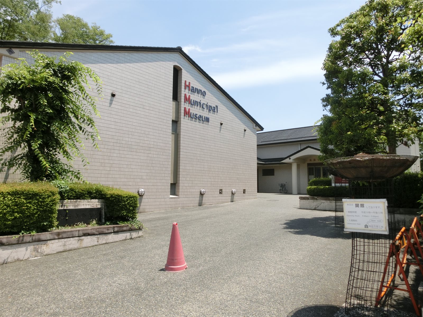 飯能市立博物館の外観。白いタイル張りの切妻の建物の壁に「Hanno Municipal Museum」の文字があり、右手に瓦屋根の入口棟と木立が見える