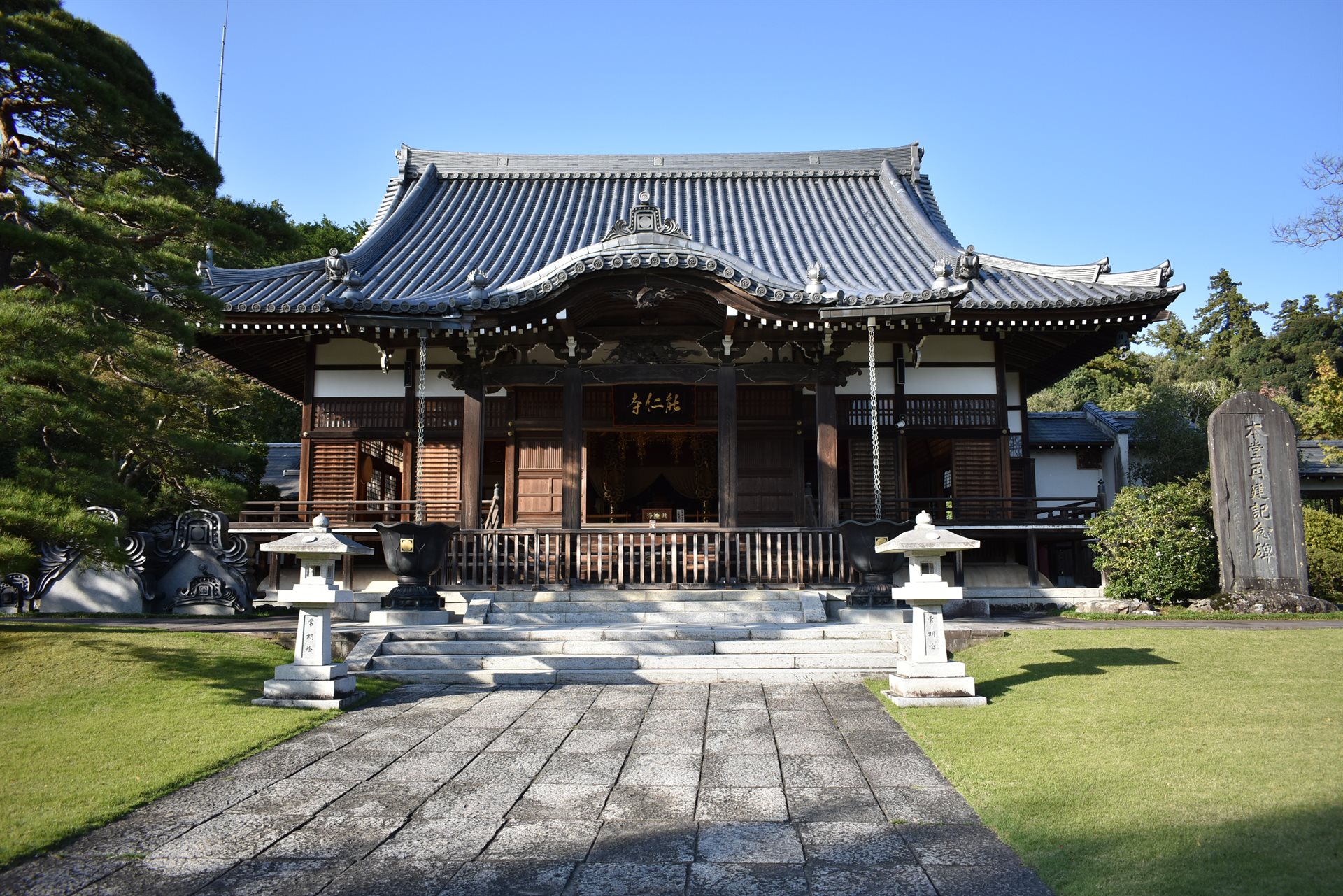 能仁寺の本堂。石畳の参道の先に、瓦屋根と木の扉の本堂が建ち、両側に芝生と石灯籠がある。背景は青空