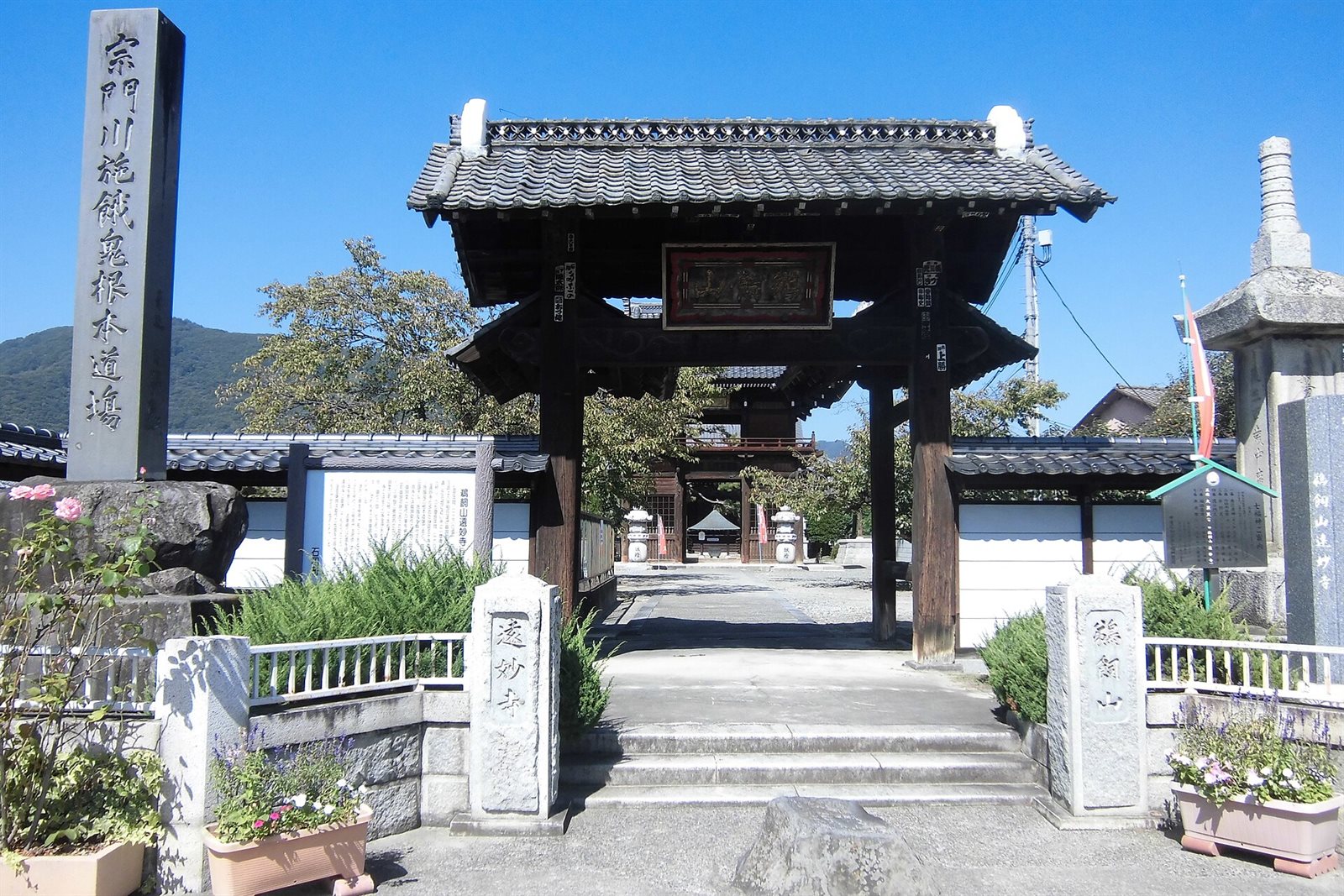 遠妙寺の山門。瓦屋根の木造の門の左に「宗門川施餓鬼根本道場」と刻まれた石柱、門前の石柱に「遠妙寺」「鵜飼山」の文字があり、奥にもう一つの門が見える
