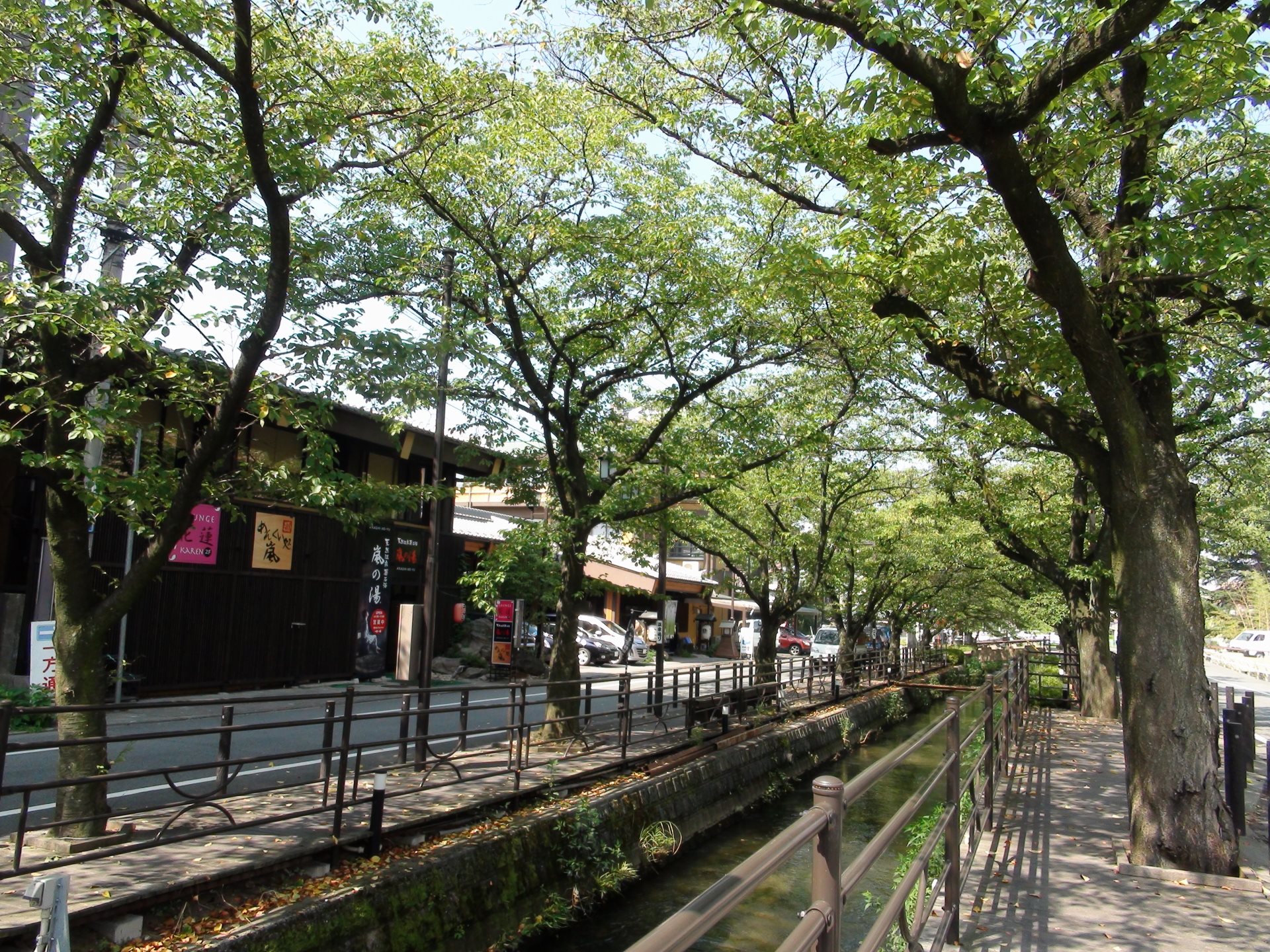 石和温泉の温泉街。小川沿いに柵のついた遊歩道と並木が続き、左手に旅館や店の建物が並んでいる