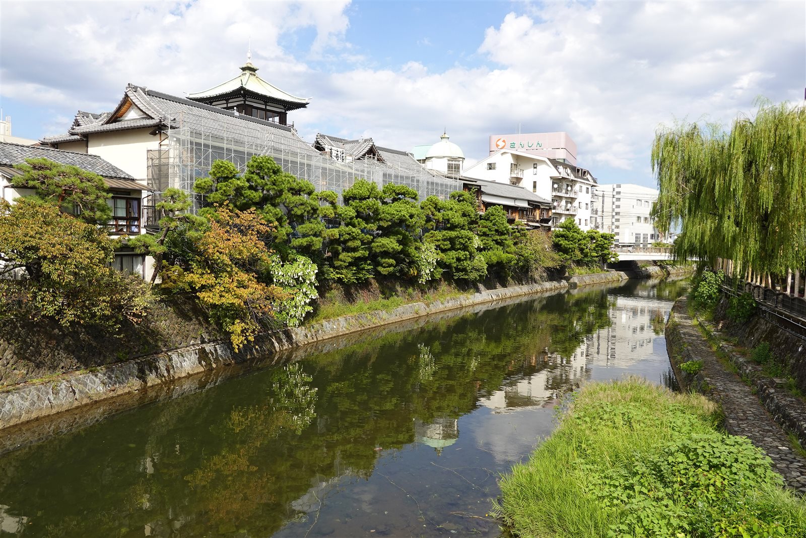 晴れた空の下、松川沿いに建つ木造三階建ての東海館と、川面に映る柳と温泉街の建物