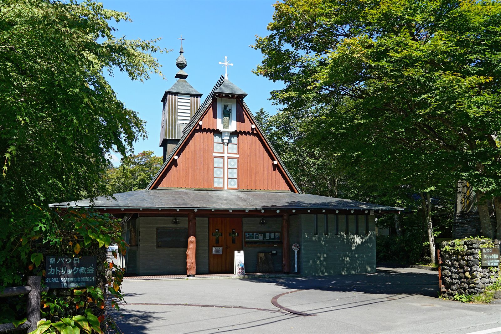 緑の木々に囲まれた聖パウロカトリック教会の正面。三角屋根の上にマリア像、左奥に鐘楼が立つ