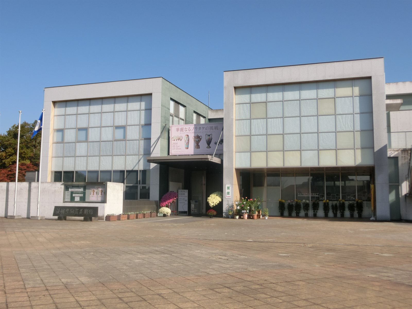 茨城県陶芸美術館の外観。四角い窓の並ぶ白い建物が左右に建ち、中央の入口に展覧会の垂れ幕が下がる。入口の前に鉢植えの菊が並んでいる