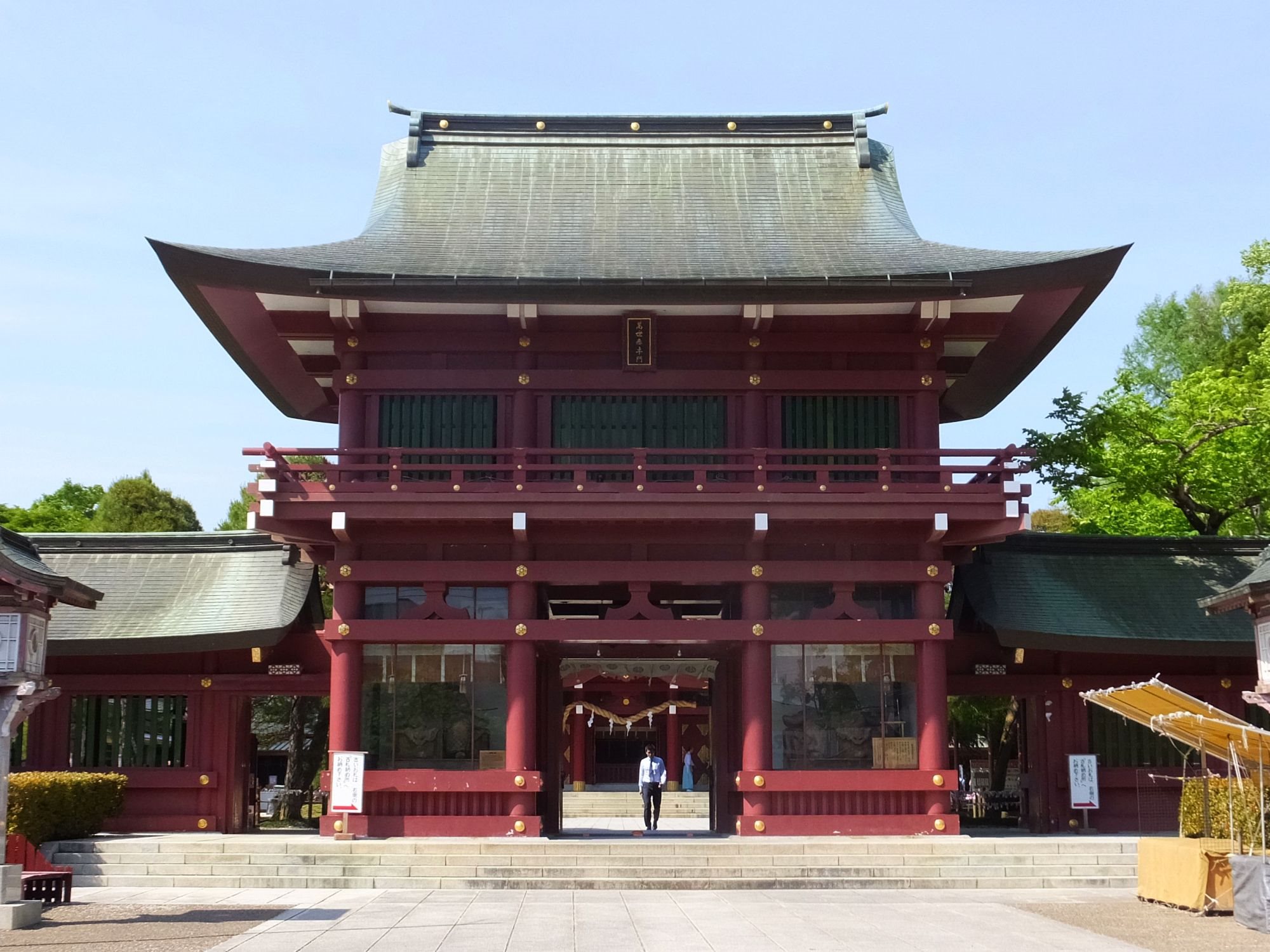 笠間稲荷神社の楼門。朱塗りの二層の門が正面に立ち、左右に塀が続く。手前に石畳の参道と階段、背景は青空