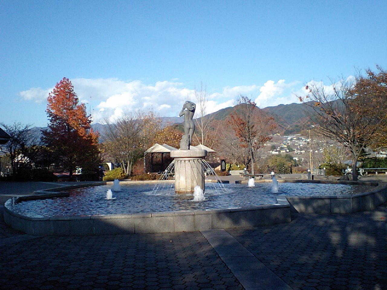 ぶどうの丘の噴水広場。石畳の広場の中央に円形の水盤と彫像のある噴水があり、まわりに紅葉した木が立つ。奥に町並みと山並み、青空が見える