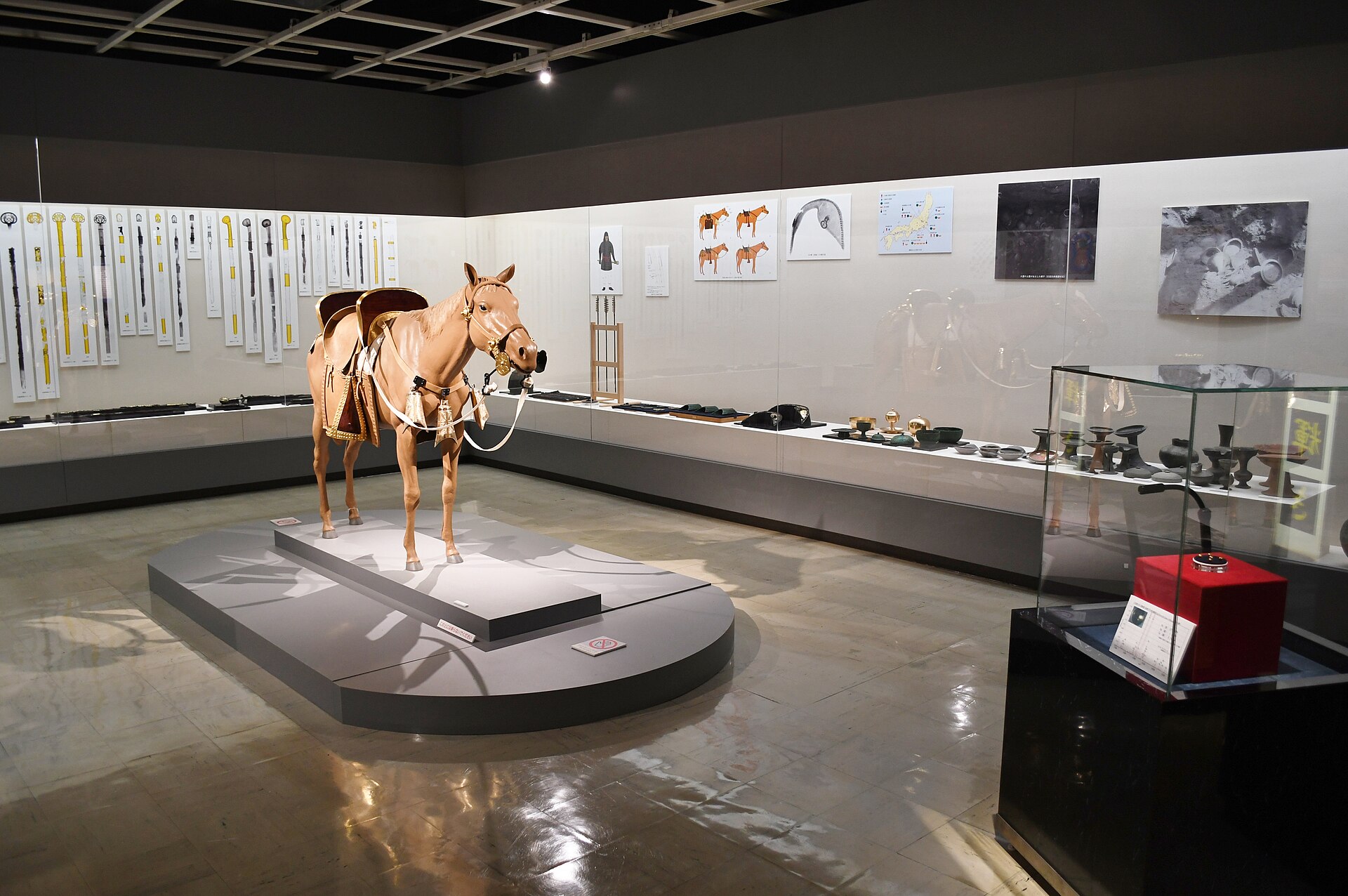 馬の模型と大刀の展示パネル、ガラスケースの出土品が並ぶ博物館の展示室