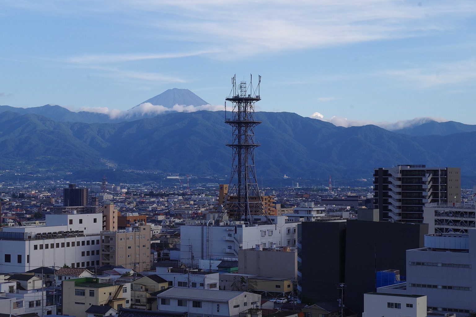 甲府の市街地の向こうに、山並みと雲をかぶった富士山が見える眺め