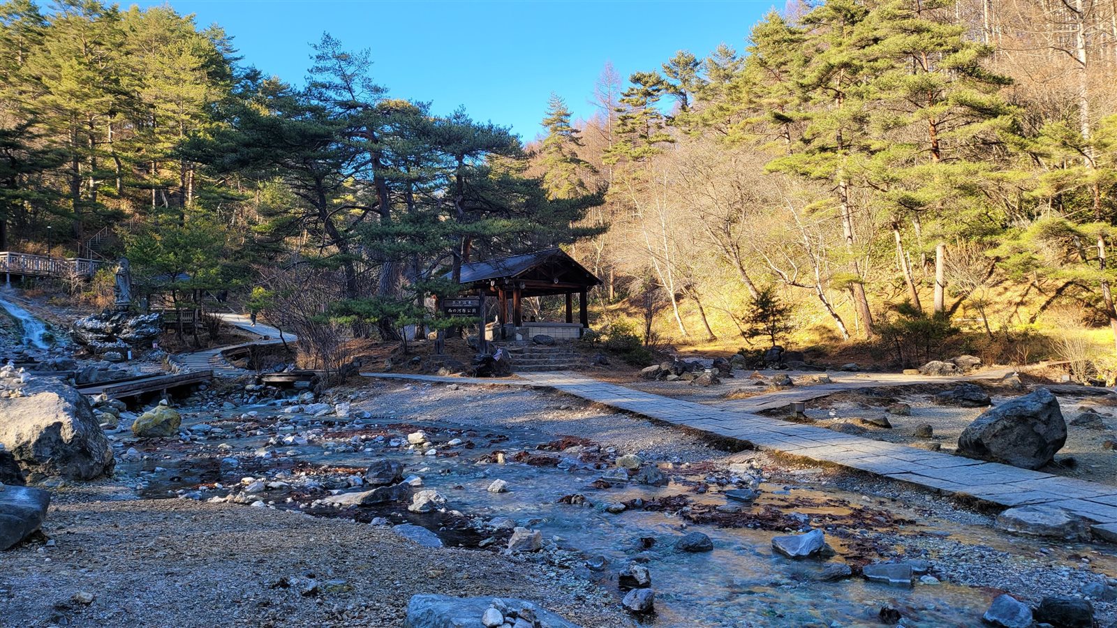 晴れた日の西の河原公園。湯の流れる河原に木道が続き、奥に東屋と松の木が見える