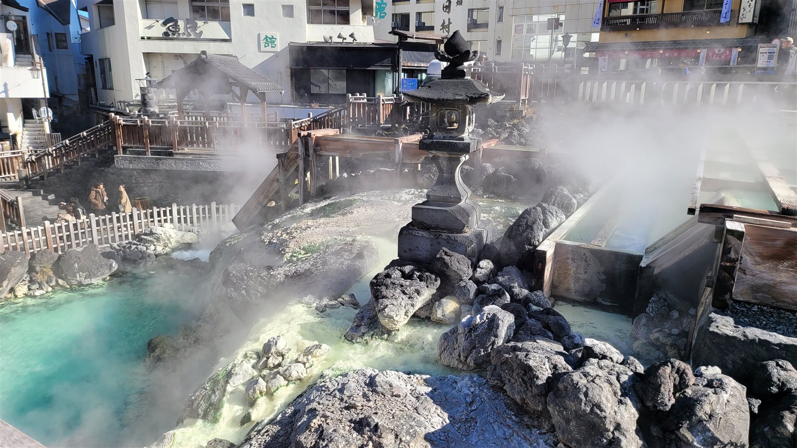 湯けむりの中、青緑色の湯がたまる湯畑の湯滝の下と石灯籠、奥に湯樋と温泉街の建物