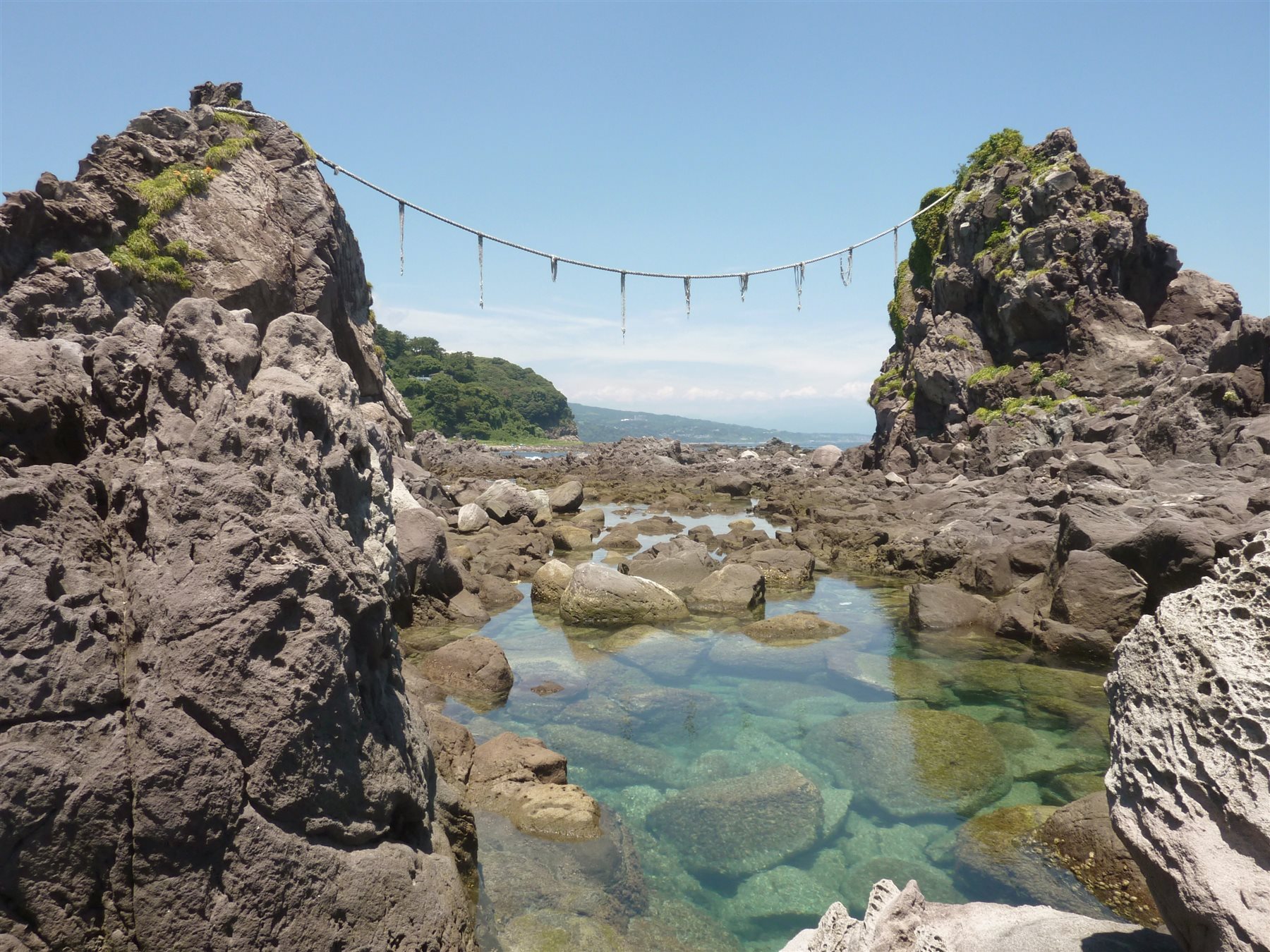 海の中に二つの大きな岩が立ち、岩の間に注連縄が張られ、手前に青緑色の潮だまりが広がる三ツ石