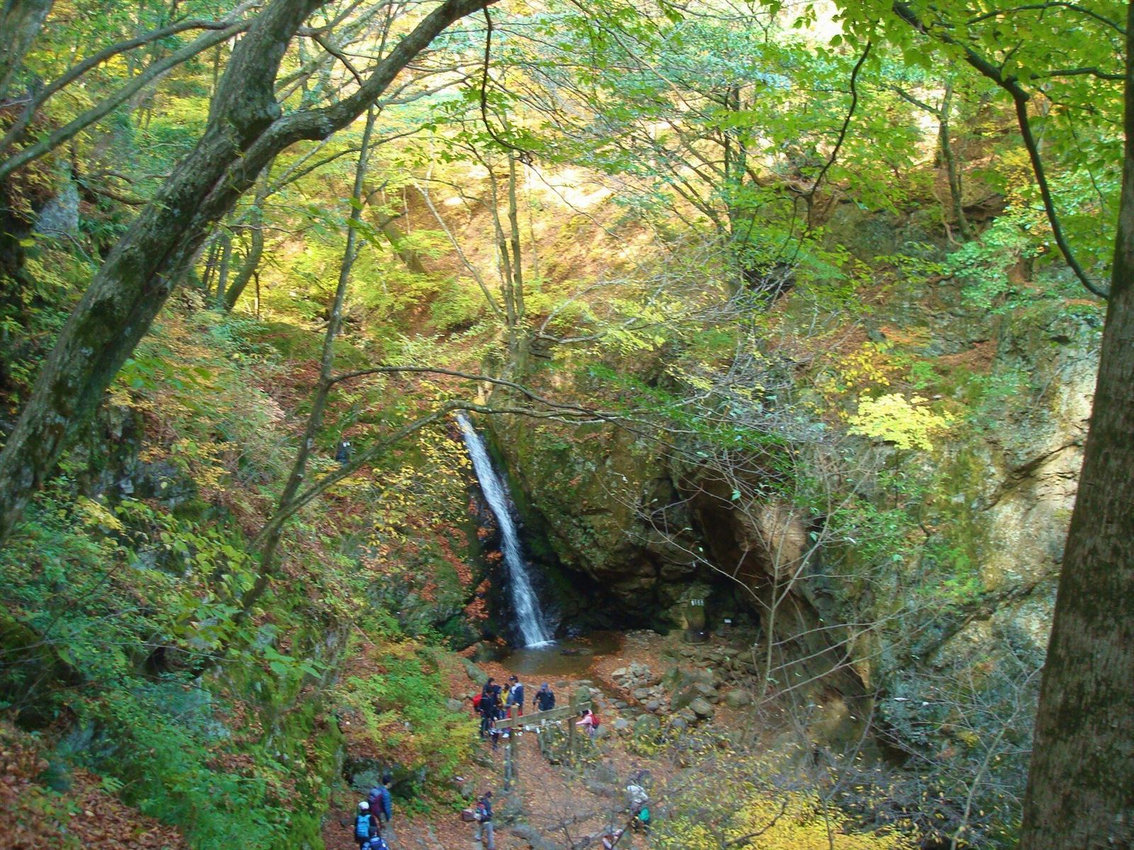 紅葉の木々の谷あいに、細い滝が落ちる綾広の滝と、歩く登山者たち