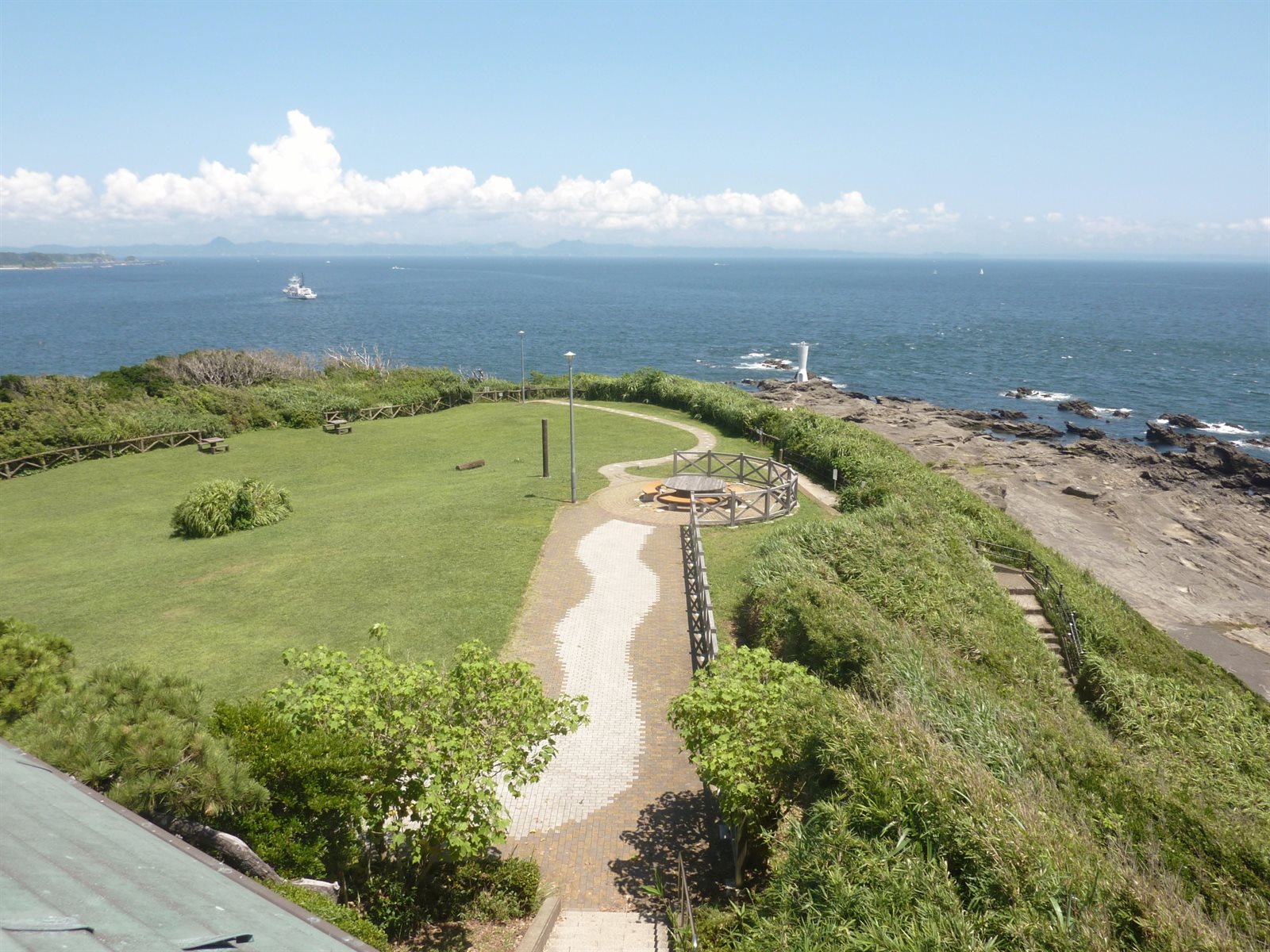 城ヶ島公園の展望台から見た景色。芝生の広場と遊歩道の先に岩場があり、その向こうに青い海と水平線、遠くの陸地が見えている