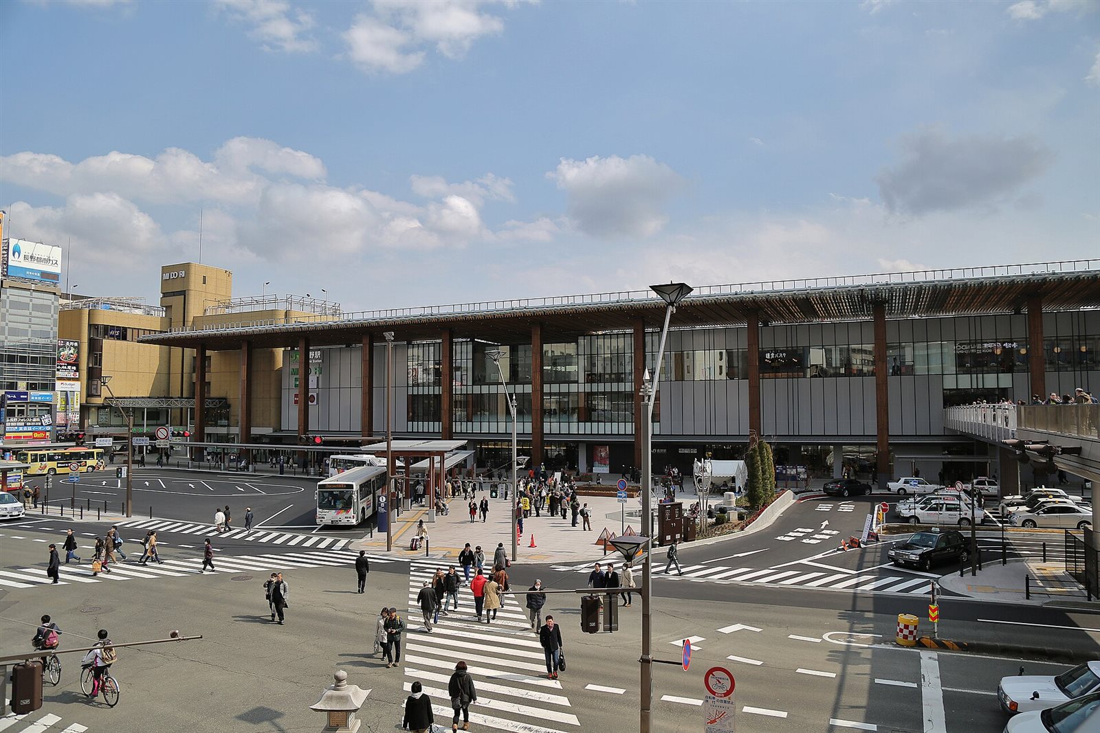 晴れた空の下、木の柱が並ぶ大きなひさしのある長野駅善光寺口と、駅前の横断歩道