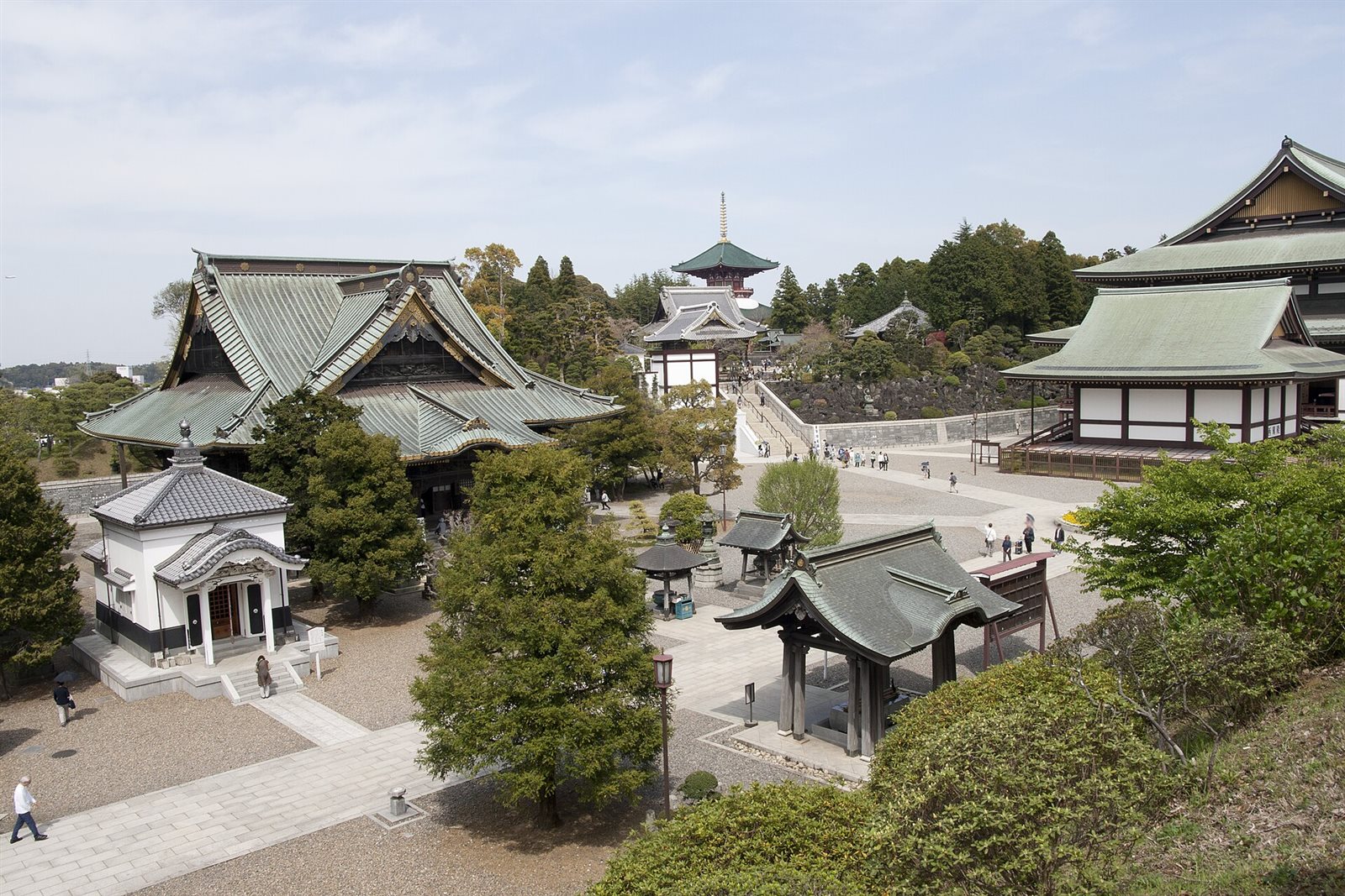 高い位置から見た成田山新勝寺の境内。緑青色の屋根を持つお堂がいくつも並び、奥の木立の中に塔が見え、参道を参拝者が歩いている