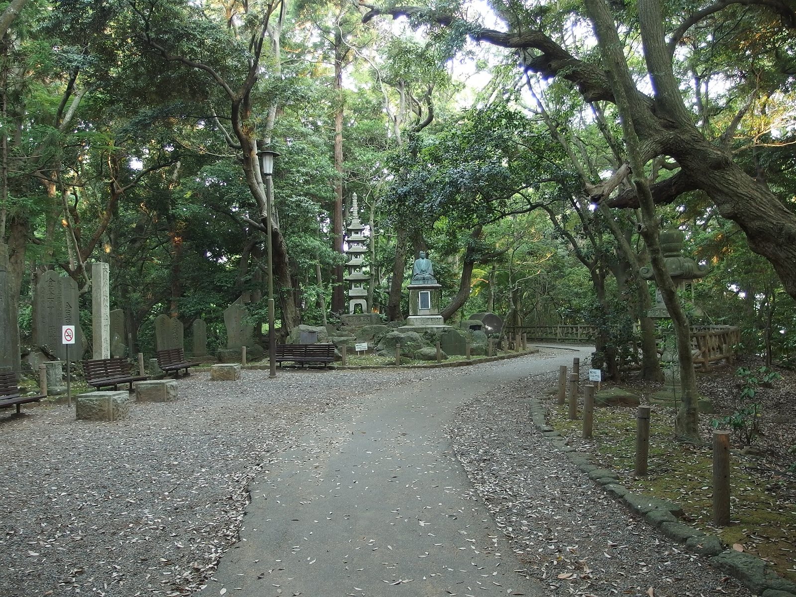 成田山公園の散策路。大きな木々に囲まれた砂利混じりの道が奥へ続き、脇にベンチと石碑、石塔が並んでいる