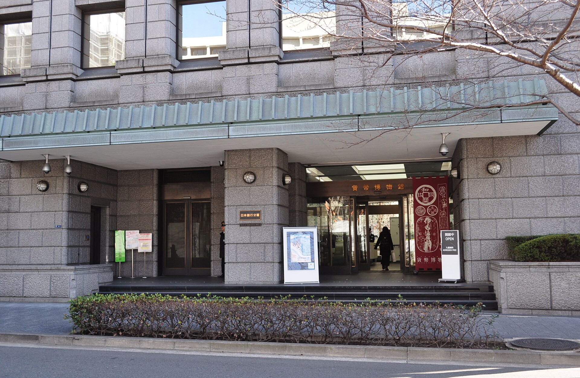 石張りの外壁の建物の入口に「貨幣博物館」の看板が見え、手前に生け垣と、赤いのぼりが立つ