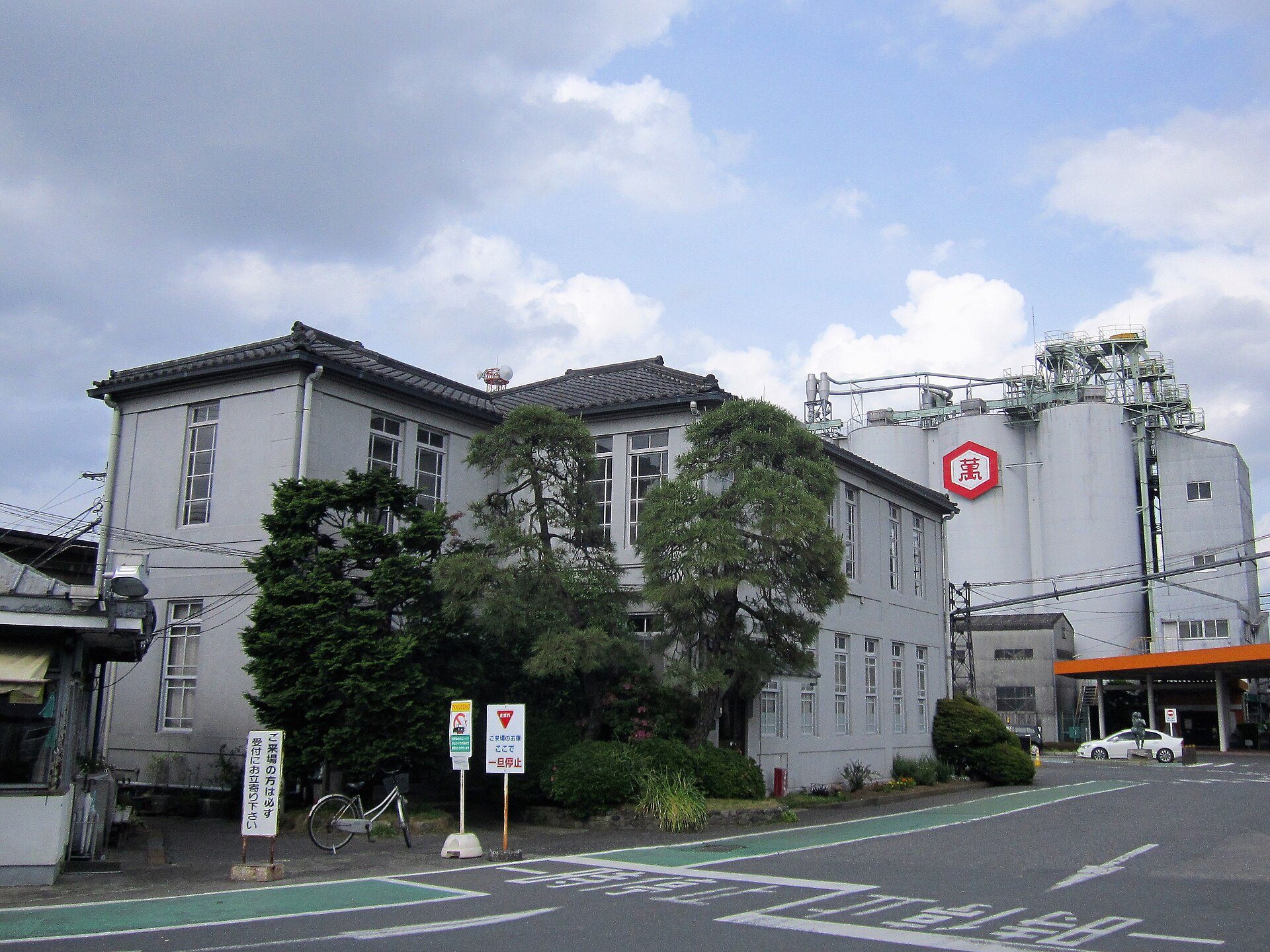 灰色の壁の二階建ての古い事務所の建物と、その奥に立つ「萬」の印が付いたしょうゆの貯蔵タンク