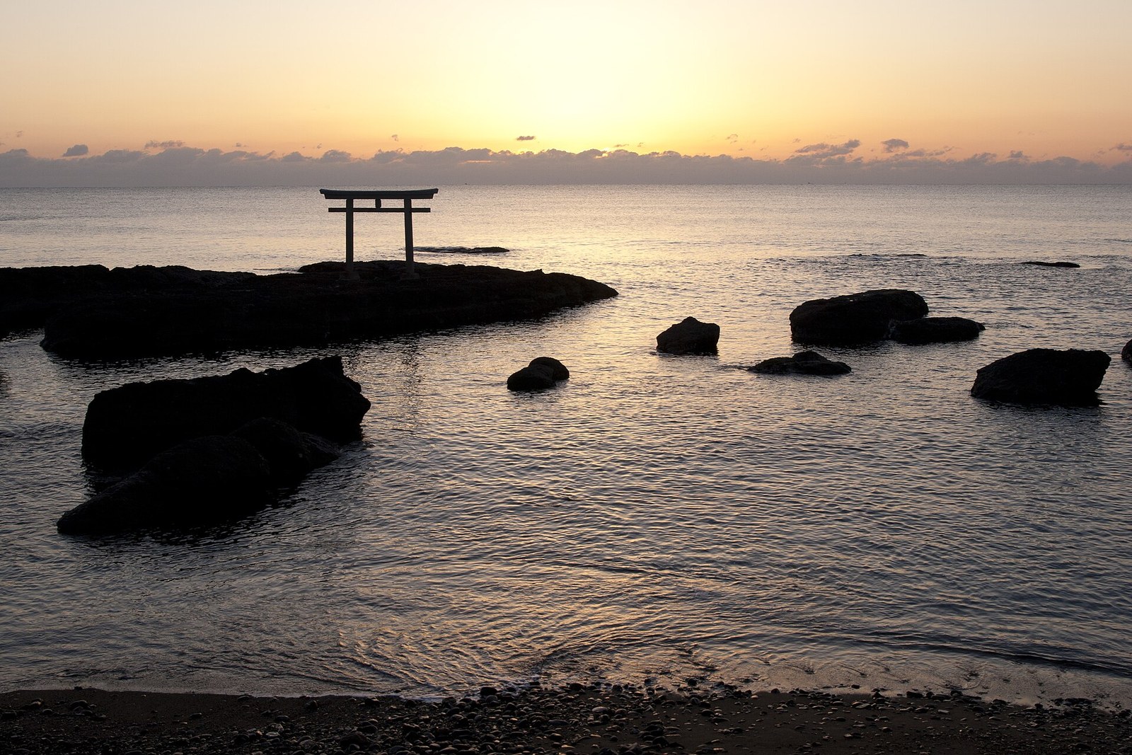 朝焼けの海に浮かぶ神磯の鳥居のシルエット。手前は海岸の小石