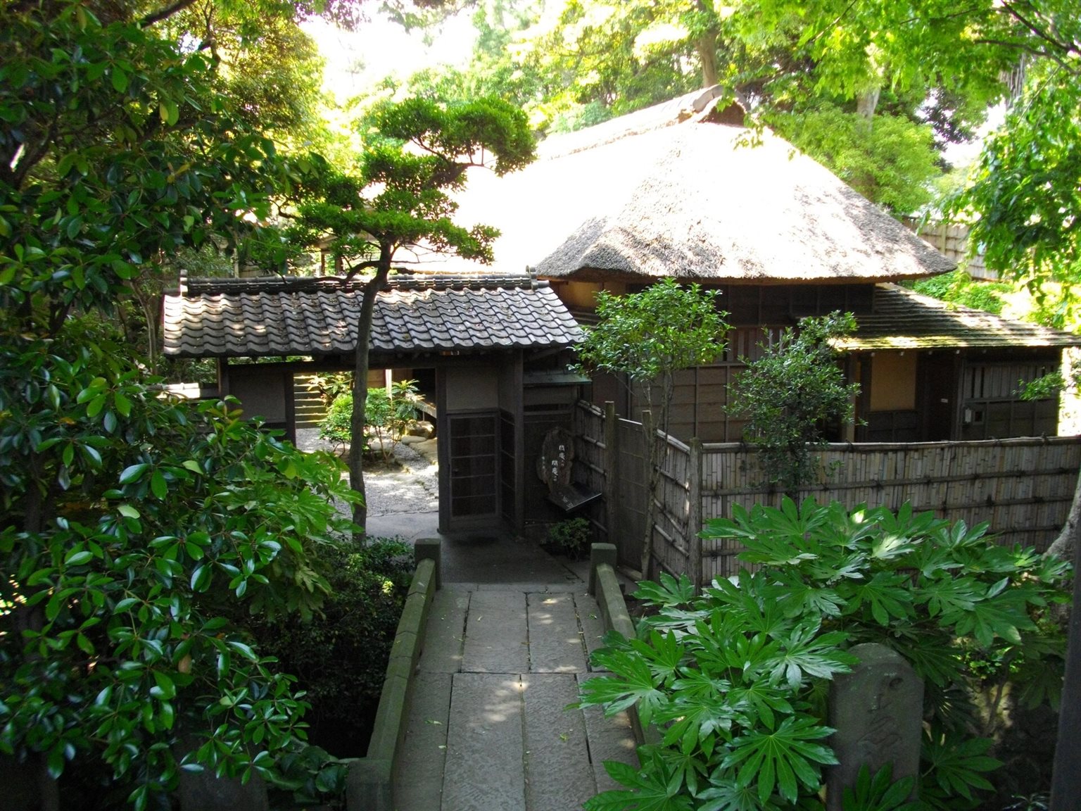 木々に囲まれた石畳の先に、茅葺き屋根の草庵と瓦屋根の門が見える鴫立庵