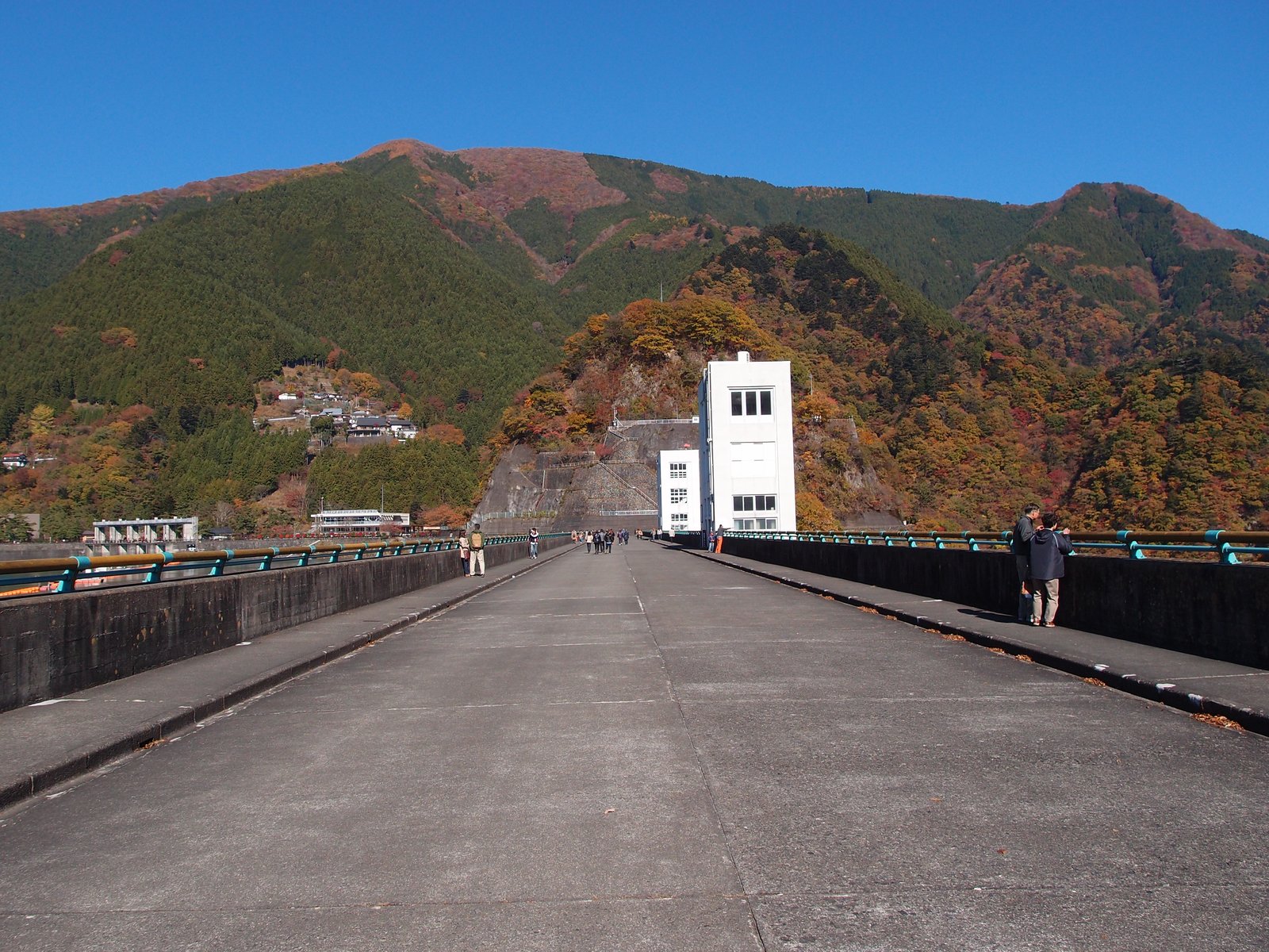 小河内ダムの堤頂の道。左右に低い欄干が続くまっすぐなコンクリートの道の先に白い塔の建物が立ち、奥には紅葉の混じる山がそびえる。道の上を何人かが歩いている