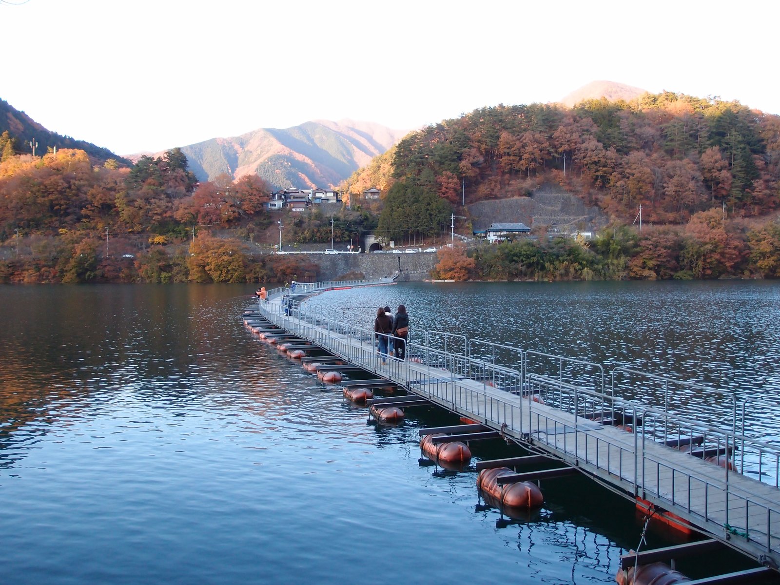 奥多摩湖の麦山の浮橋。赤い浮きの上に手すりつきの細い通路が湖面を横切って続き、2人が歩いている。対岸には紅葉した山と集落がある