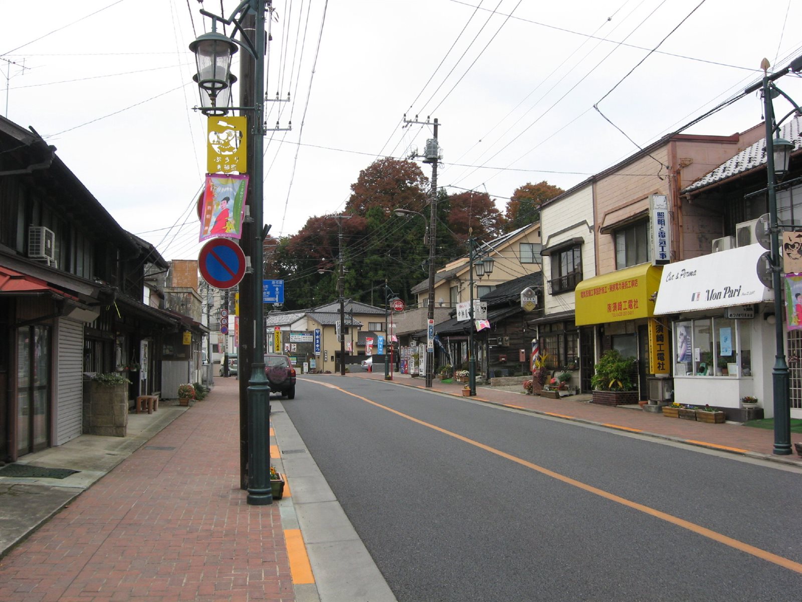 青梅市街の旧青梅街道。片側に古い木造の商家、反対側に看板を掲げた店が並び、歩道にレトロな街灯と幟が立っている