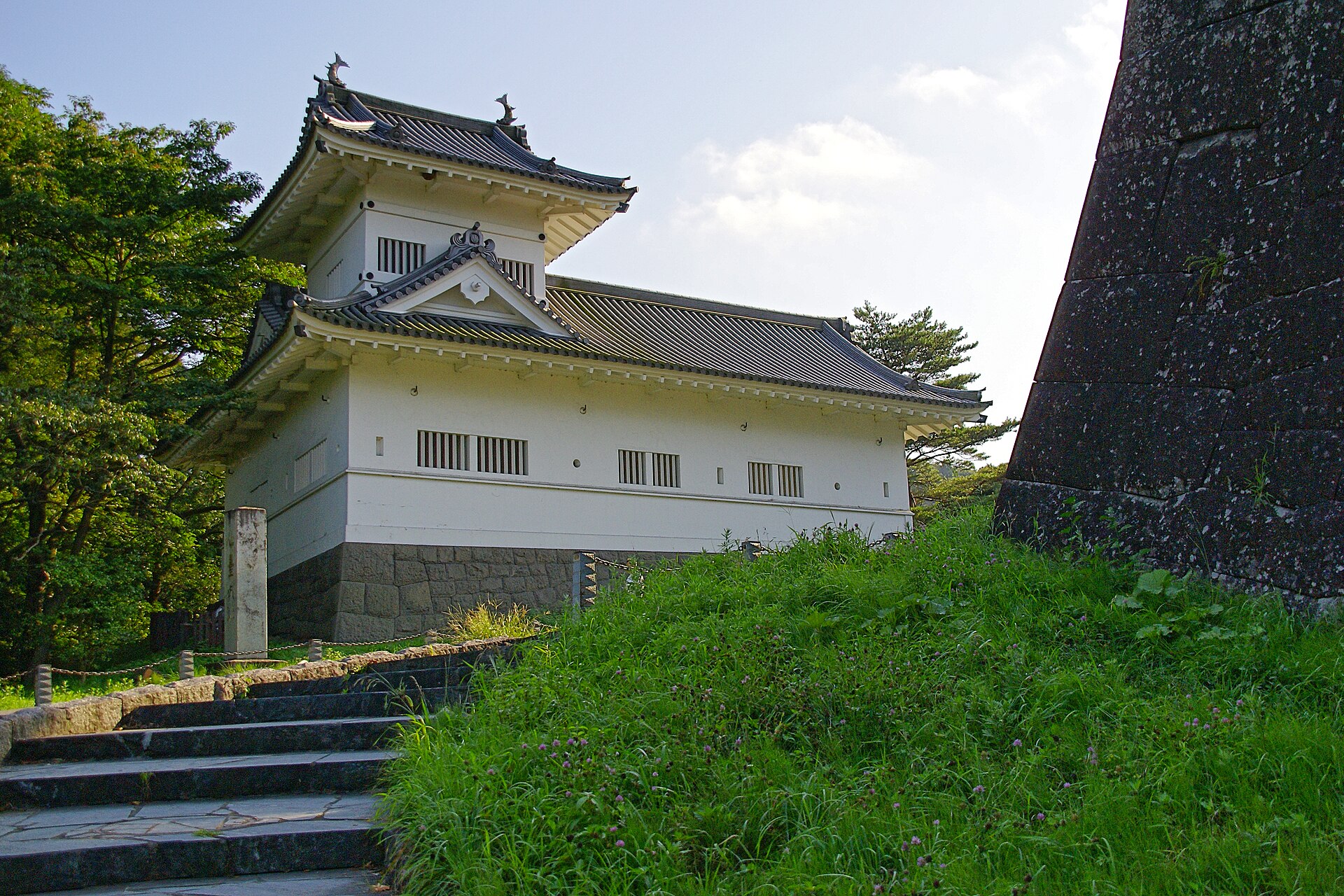 草地の斜面の上に建つ、白壁に瓦屋根の復元された脇櫓と、右に立つ黒い石垣