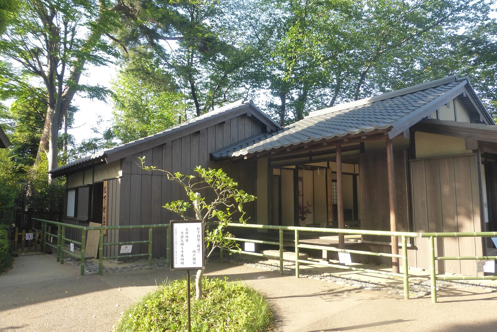木々に囲まれた、瓦屋根の平屋の建物と縁側、手前に竹の柵
