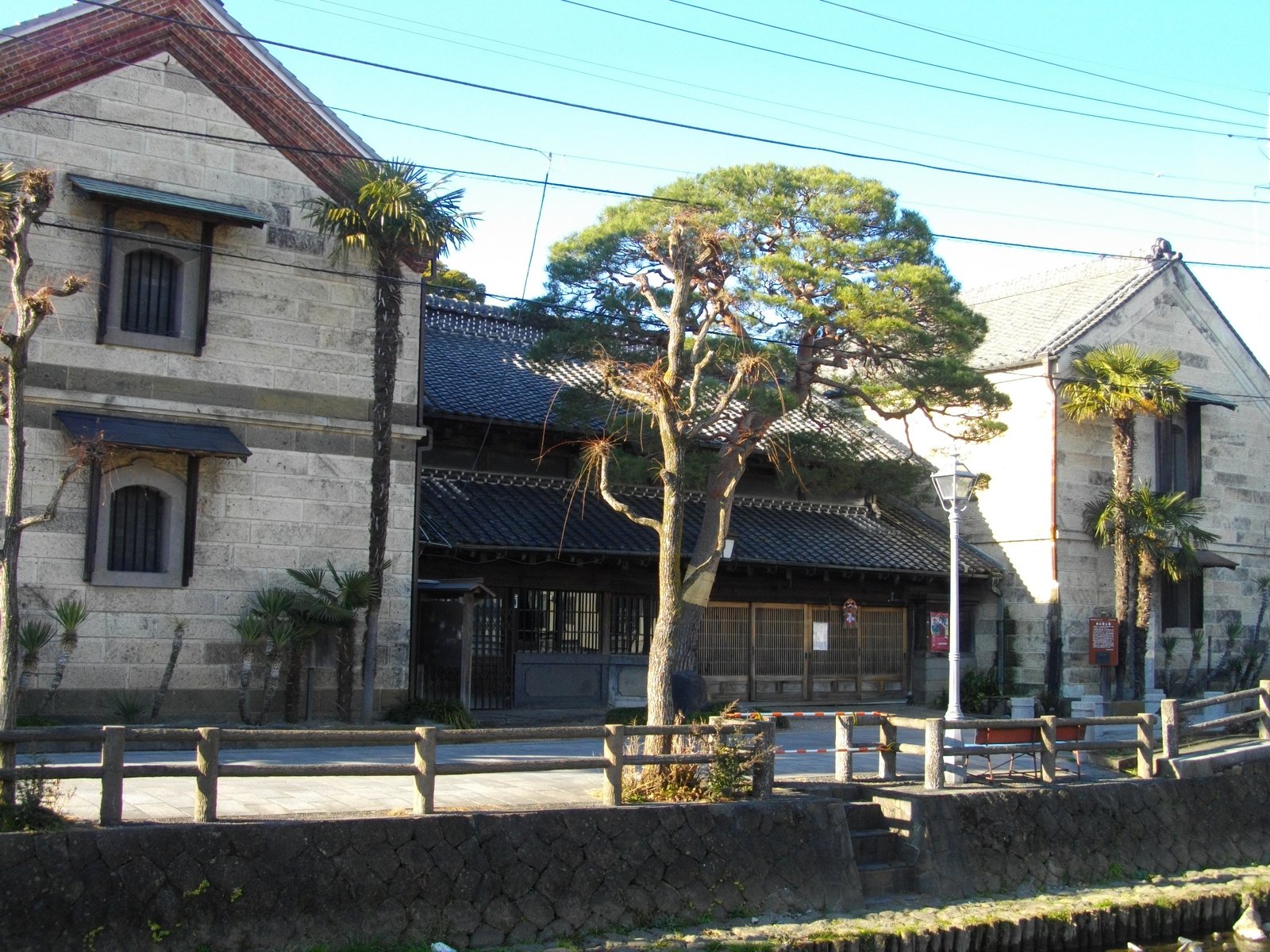 巴波川沿いの横山郷土館。瓦屋根の木造の店舗を挟んで、左右に石積みの蔵が向き合って建ち、手前に川沿いの柵と松がある