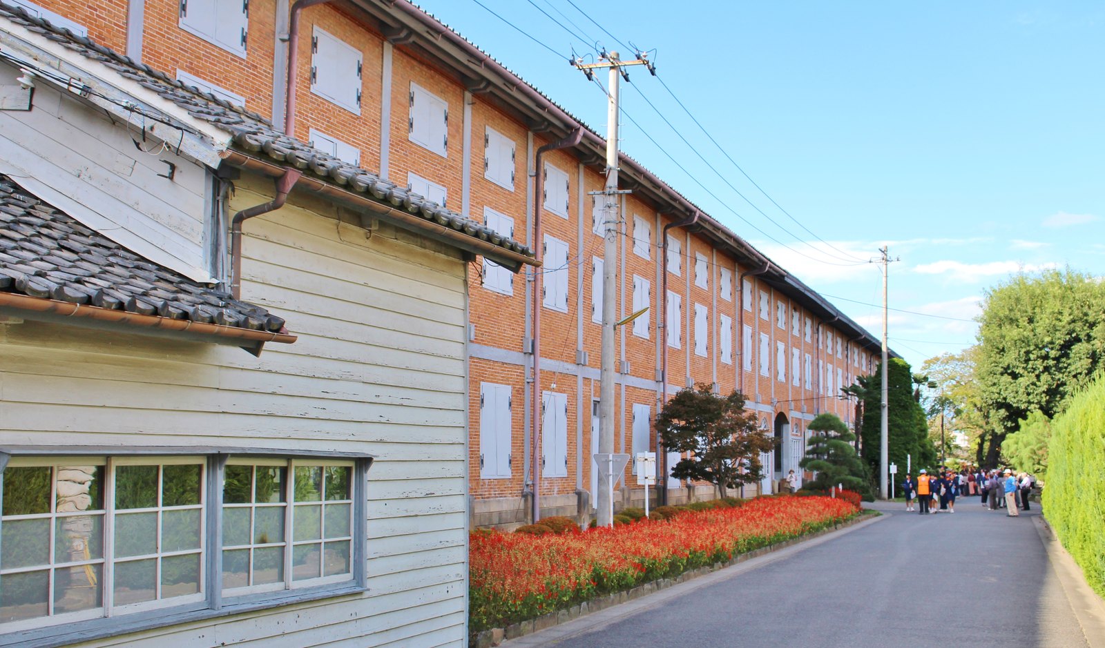 富岡製糸場の東置繭所。赤煉瓦の壁に白い窓扉が並ぶ2階建ての長い建物が奥へ続き、手前に白い板壁の木造の建物、道沿いに赤い花の花壇がある