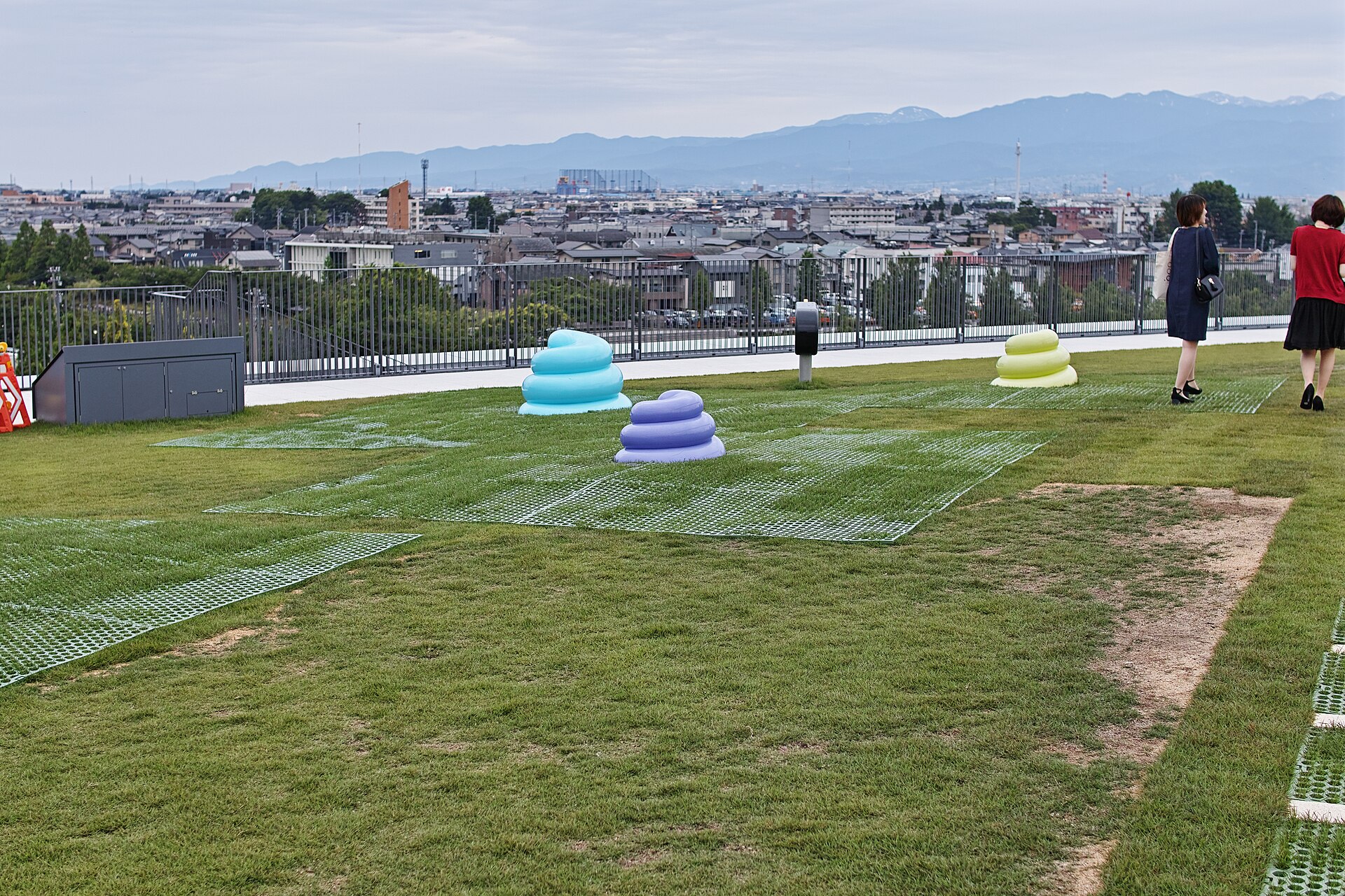 芝生の屋上に置かれた、水色・紫・黄緑の重なった形の遊具と、遠くの山並み