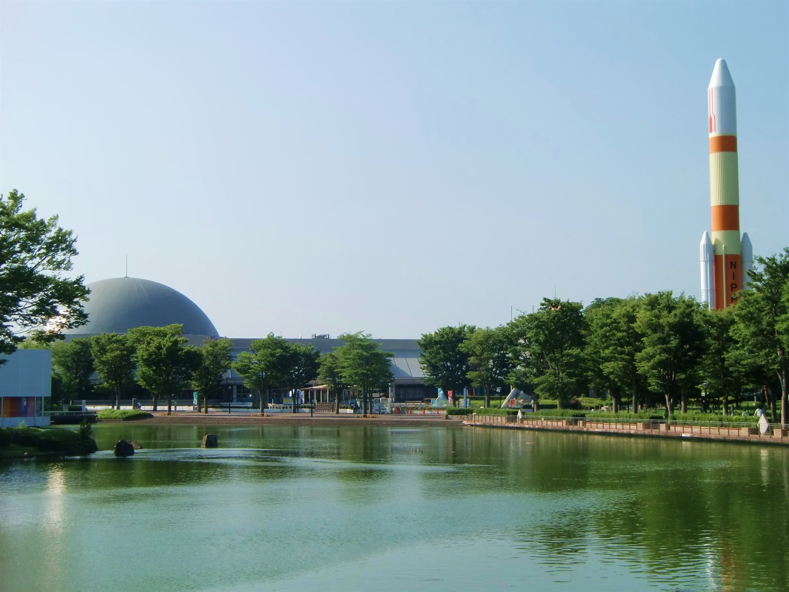 中央公園の池。緑色の水面の向こうに木立が続き、左奥にエキスポセンターの丸いドーム、右奥に木々より高くH-IIロケットの模型が見える
