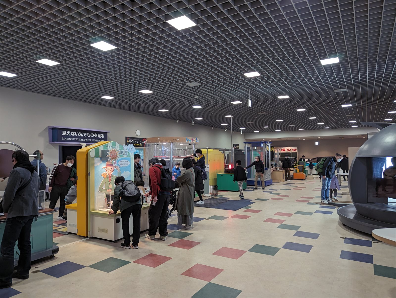 つくばエキスポセンターの展示場。格子状の天井の下に体験型の展示装置が並び、来館者が装置を囲んでのぞき込んでいる。床には色違いのひし形の模様がある