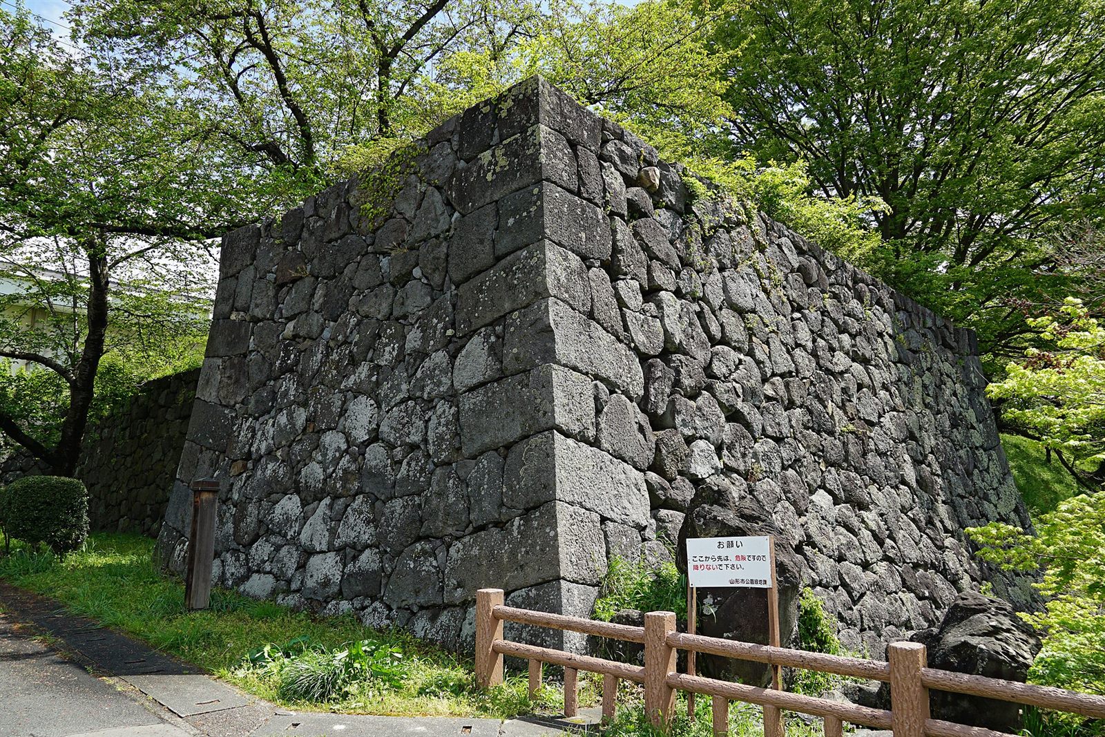 緑の木々に囲まれて、大きな石を積んだ石垣の角が立ち、手前の園路に木の柵が続いている