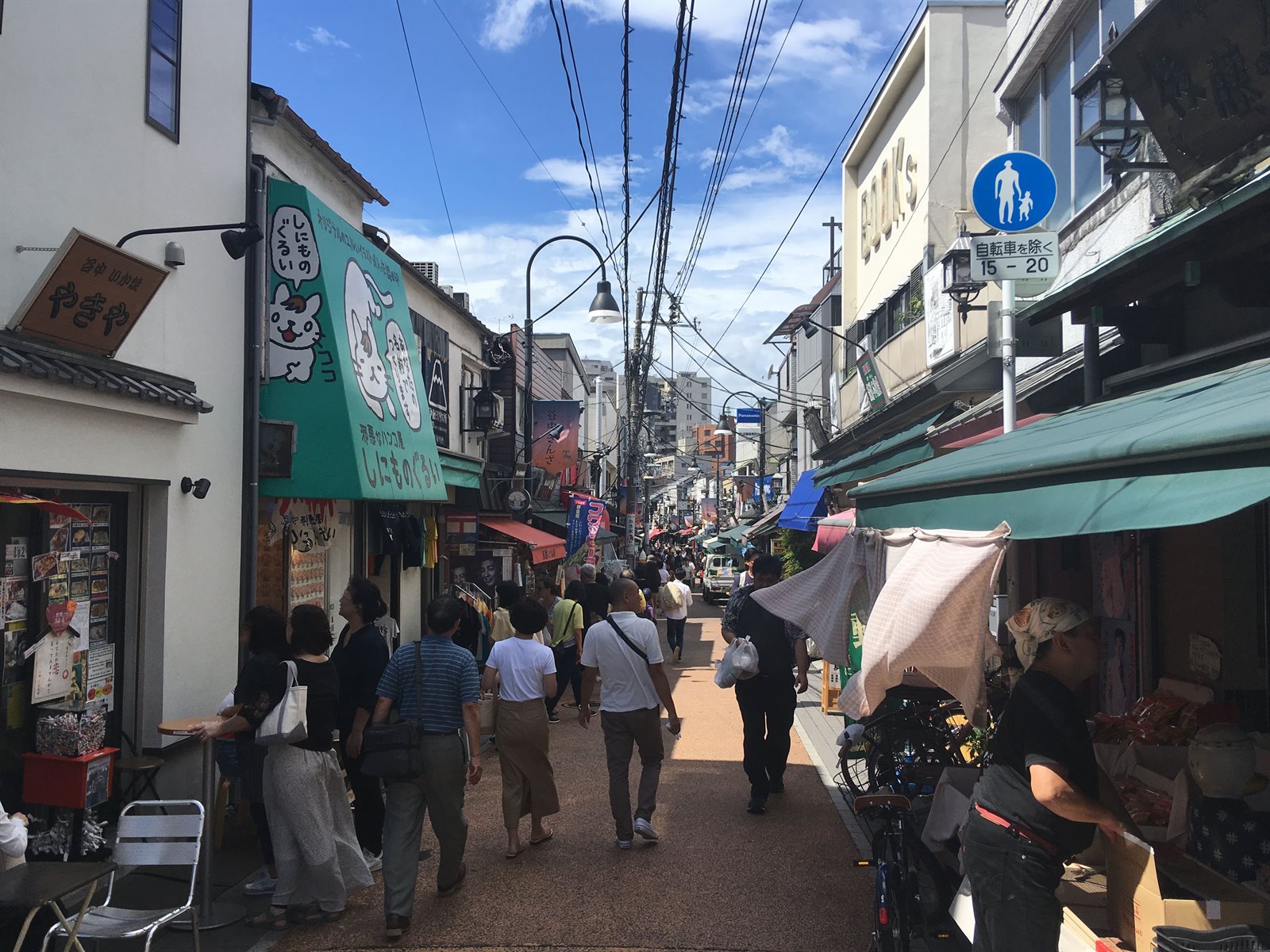 谷中銀座の通り。両側に店の看板とひさしが並び、細い道を買い物客が歩いている。頭上に電線が渡り、奥に通りが続いている