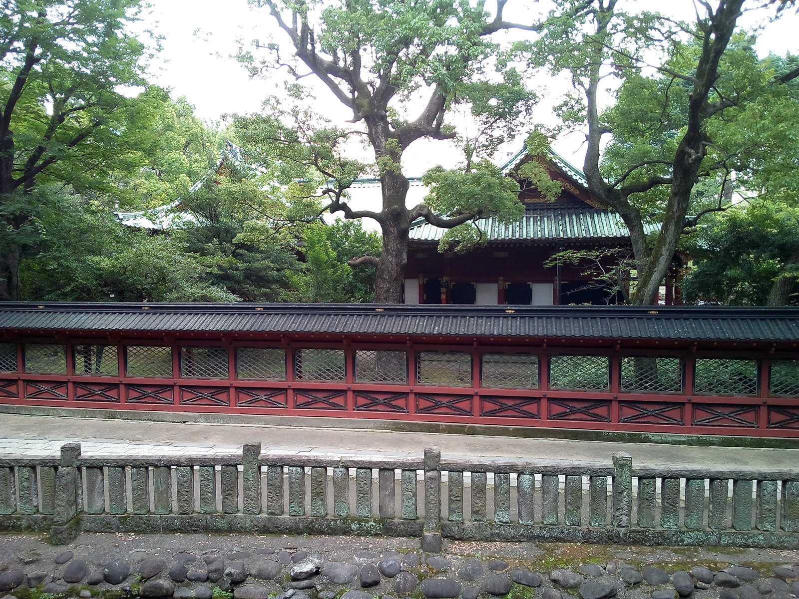 根津神社の境内。手前に石の柵、その奥に朱塗りの透塀が横に長く続き、塀の向こうに銅板葺きの社殿の屋根と大きな木が見える
