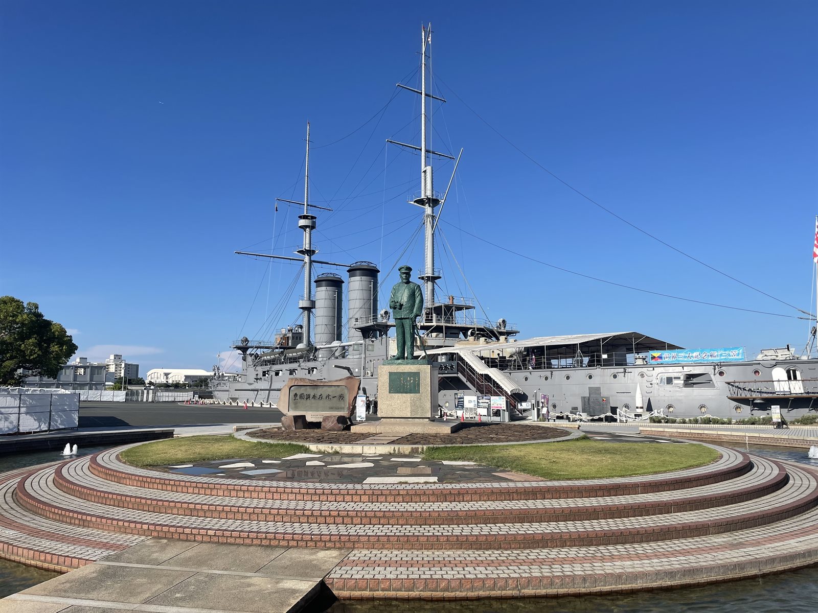 三笠公園の円形の広場に立つ東郷平八郎の像と、その奥に係留された記念艦三笠。2本のマストと2本の煙突が見え、左手に工事の仮囲いがある
