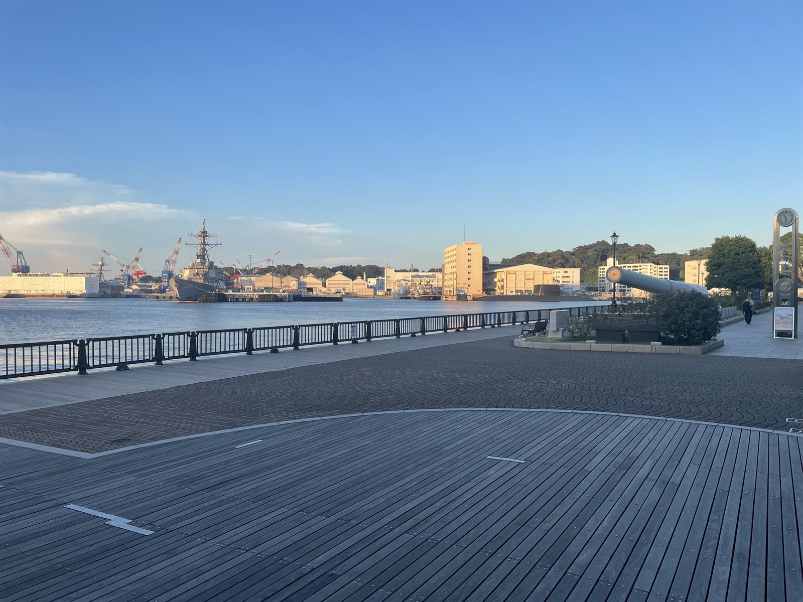 ヴェルニー公園の海沿いのボードウォーク。柵の向こうの港に灰色の艦船が停泊し、右手に戦艦「陸奥」の主砲と時計台が置かれている