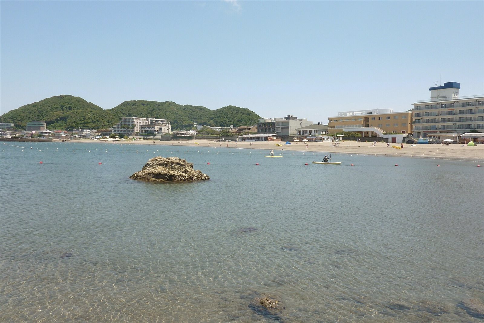浅い透明な海に岩が1つ浮かび、向こうに砂浜と建物が続く森戸海岸