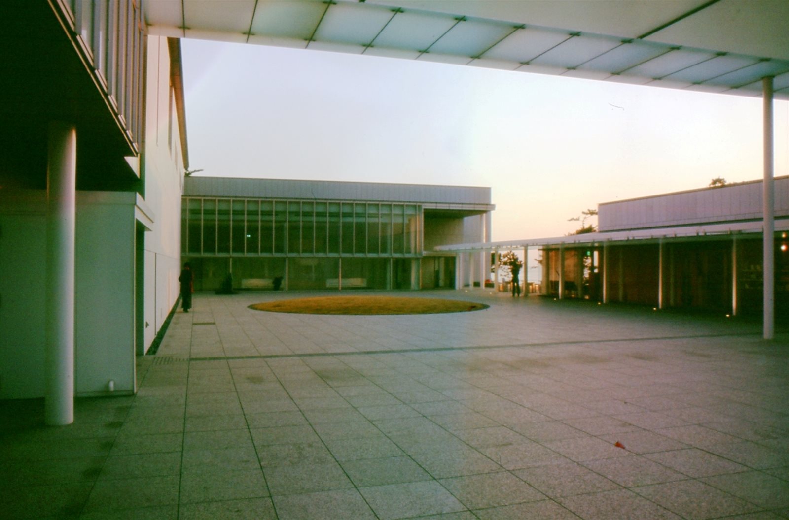 夕暮れの空の下、広い石畳の広場に面して、ガラスの壁と大きな庇のある美術館の建物が建つ